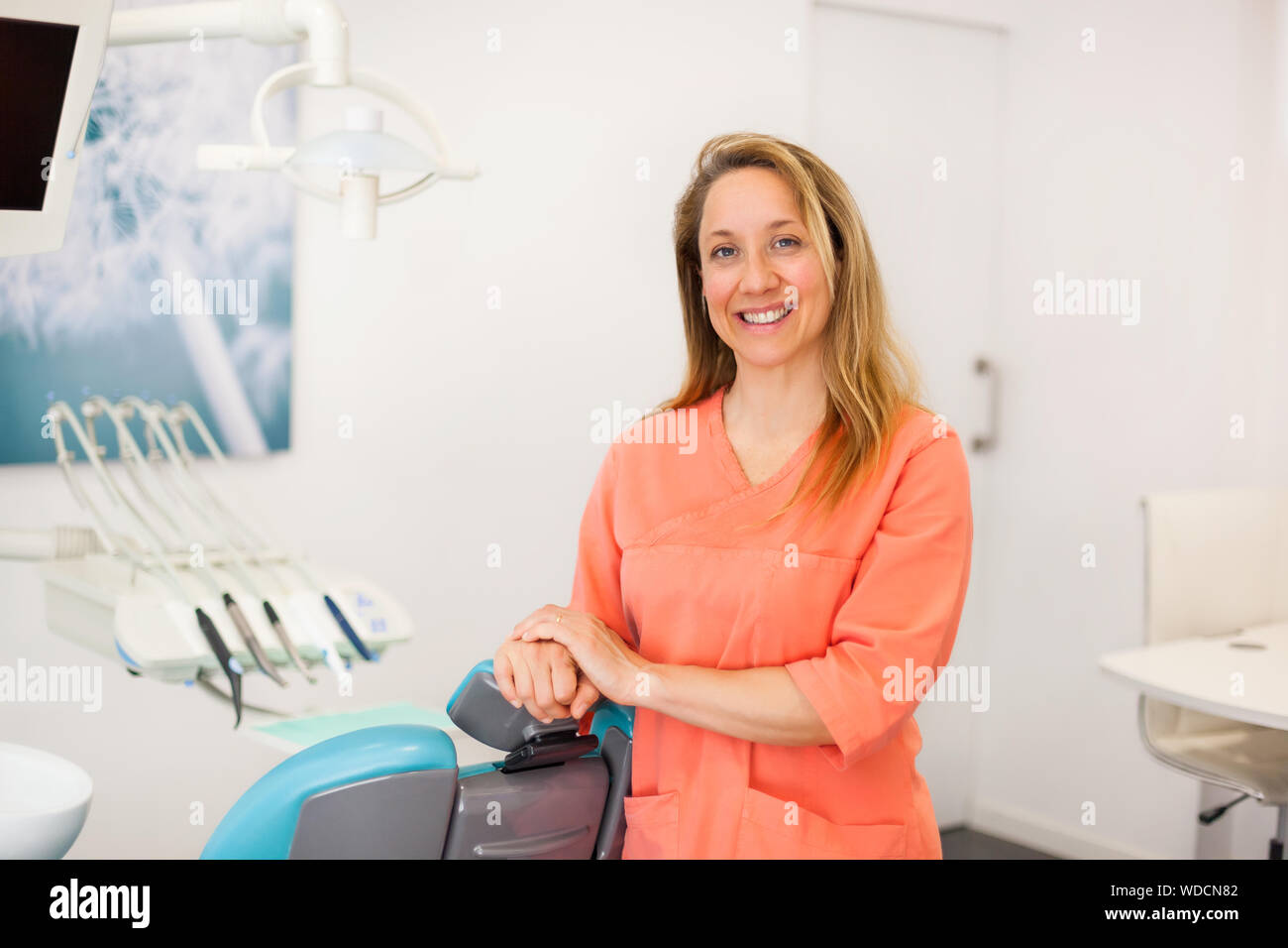 Pretty female dentist with blond hair in her surgery looking to the camera and smiling, expressing professionalism, confidence and kindness; caucasian Stock Photo