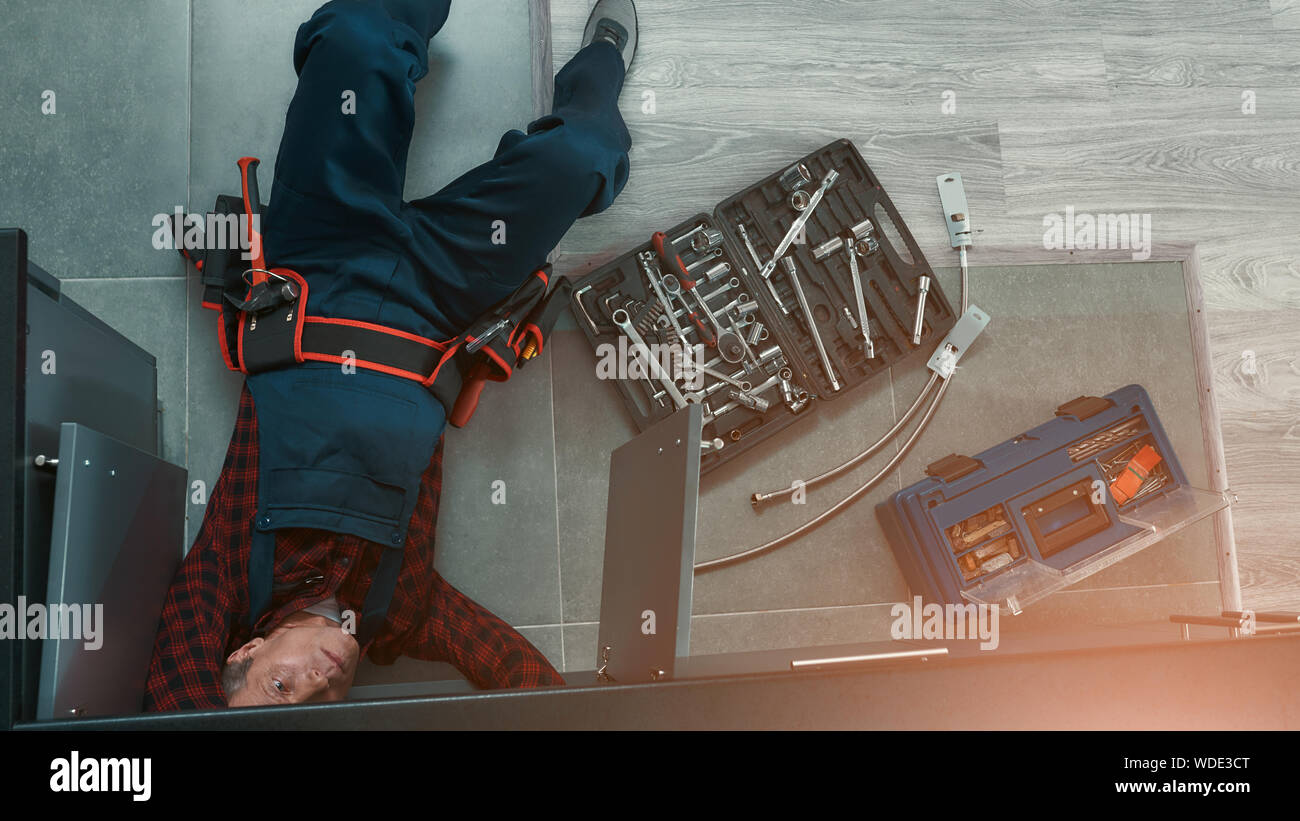 High angle view of handyman in uniform lying on floor in the kitchen and using wrench while repairing water pipe in kitchen. Toolbox on the floor. Horizontal shot Stock Photo