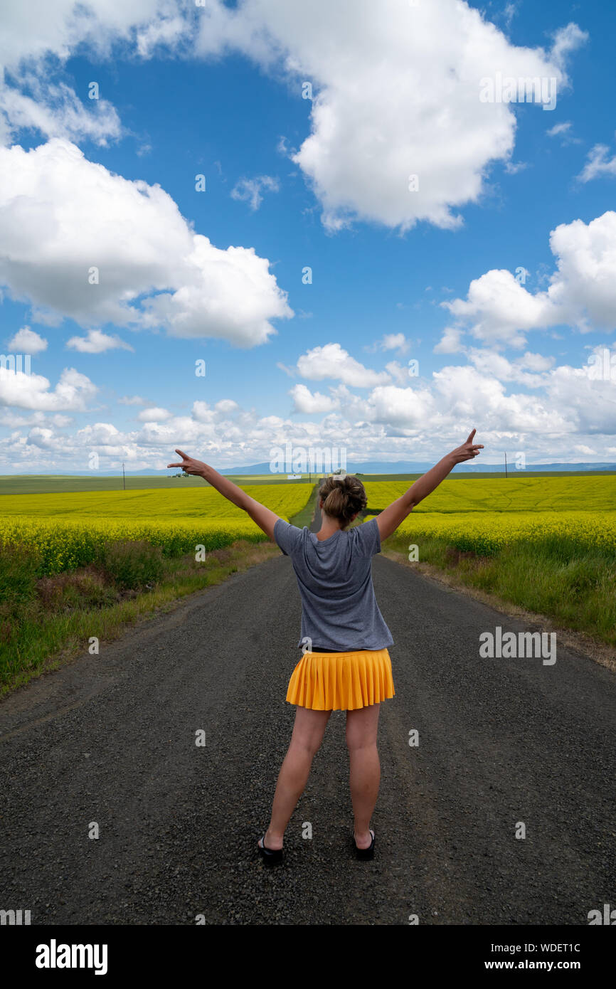 Woman with back facing camera stands in the middle of a road giving peace sign. Mustard plants on the roadside Stock Photo