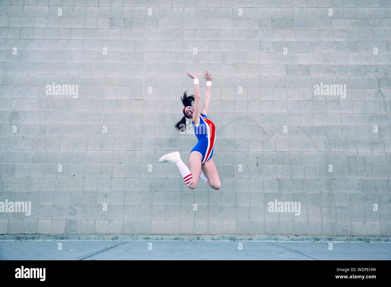 Full Length Of Cheerful Female Gymnast In Leotard Jumping Against Wall Stock Photo