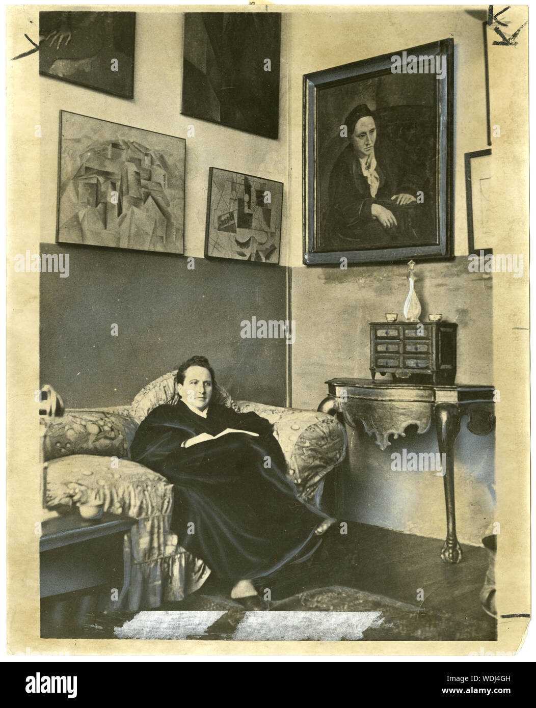 Gertrude Stein sitting on a sofa in her Paris studio, with a portrait of her by Pablo Picasso, and other modern art paintings hanging on the wall behind her Stock Photo