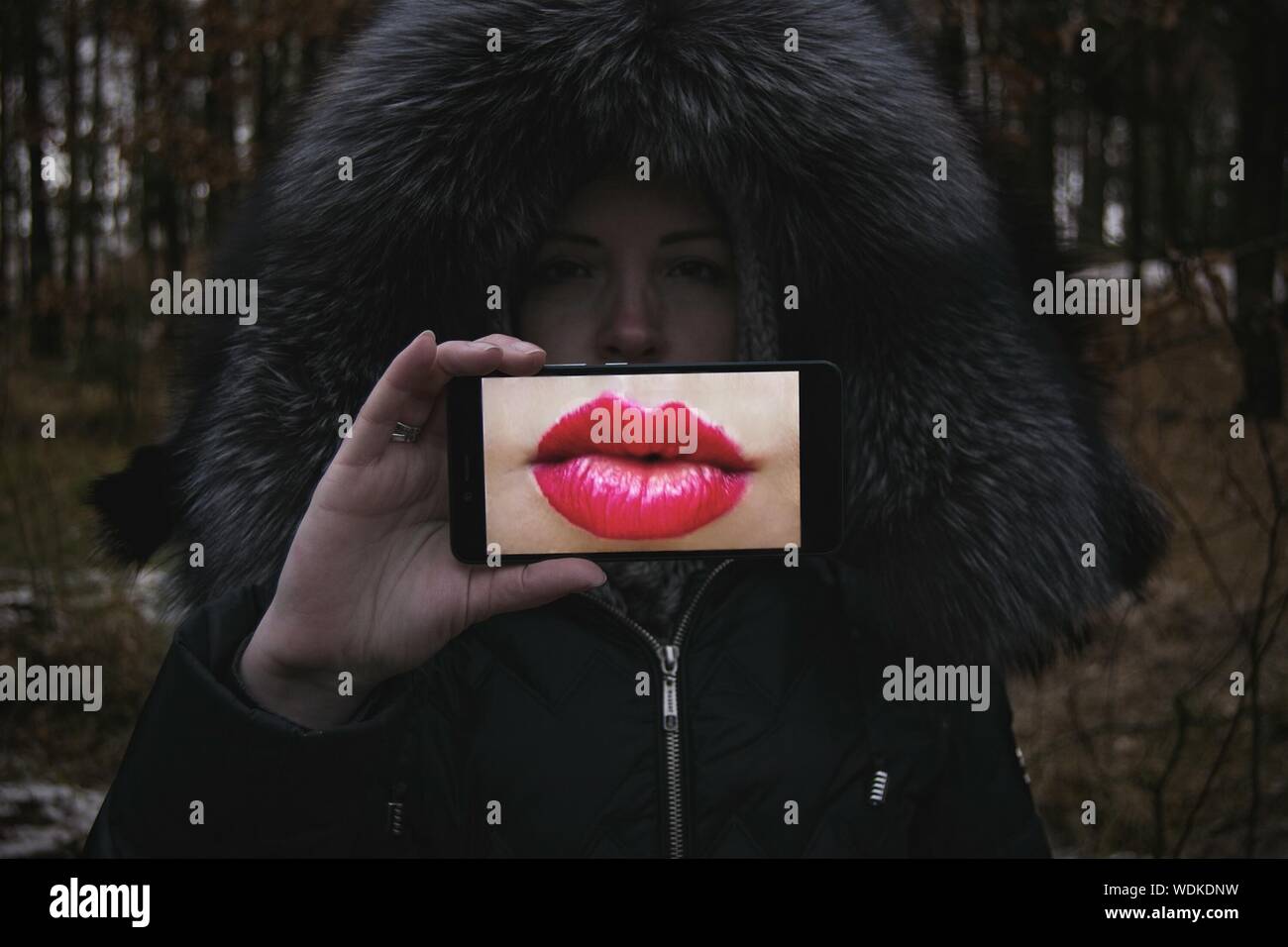 Portrait Of Young Woman Holding Smart Phone Showing Pink Lips In Display During Winter Stock Photo