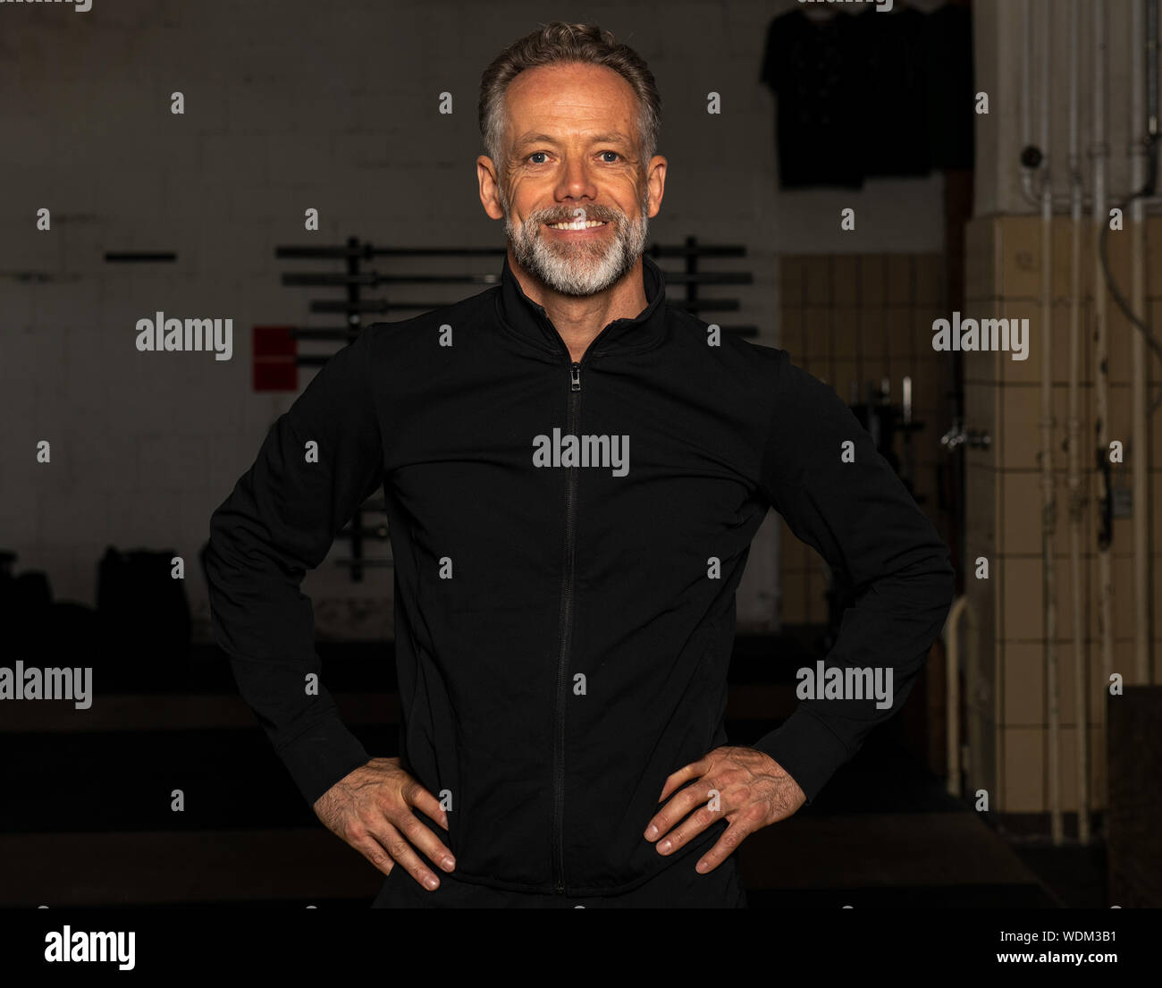Portrait of an attractive fit middle aged man in a fitness studio. Bearded grey haired man with black sweatshirt jacket is looking into the camera. Stock Photo