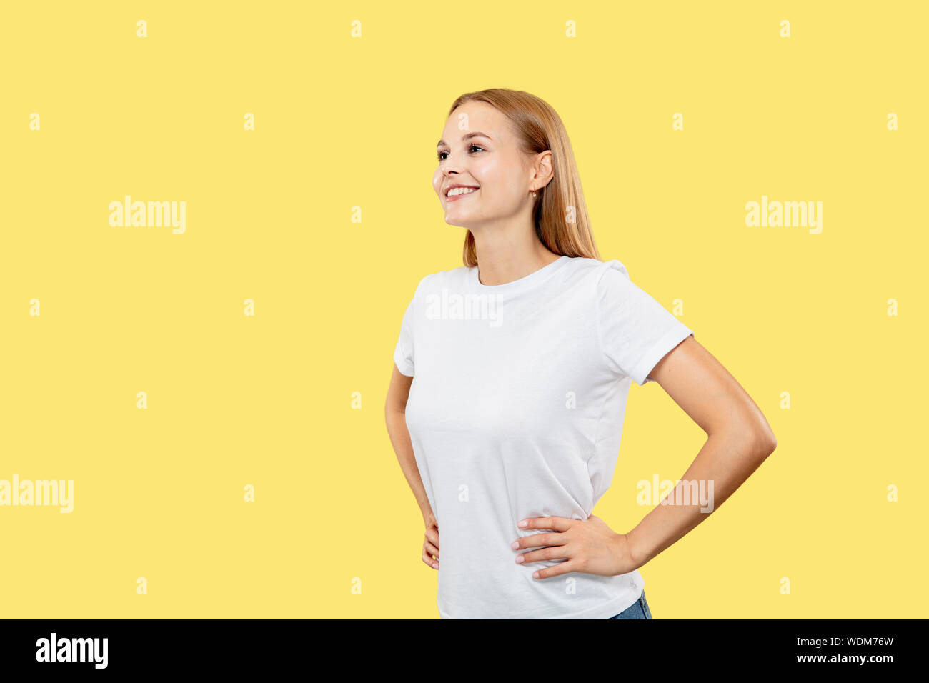 Caucasian young woman's half-length portrait on yellow studio background. Beautiful female model in white shirt. Concept of human emotions, facial expression. Smiling and standing with hands on hips. Stock Photo