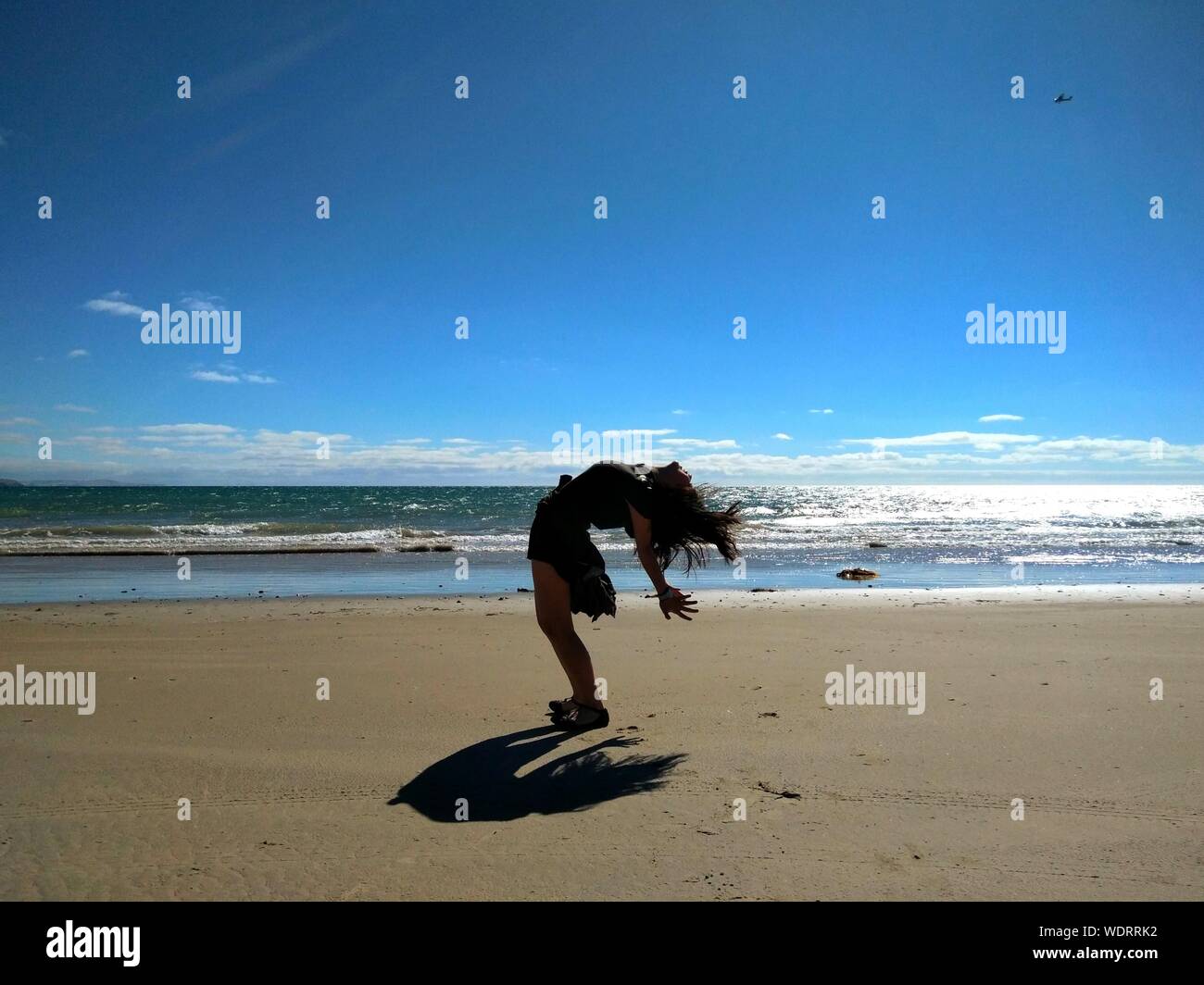 Playful Young Woman Bending Over Backwards At Beach Against Blue Sky
