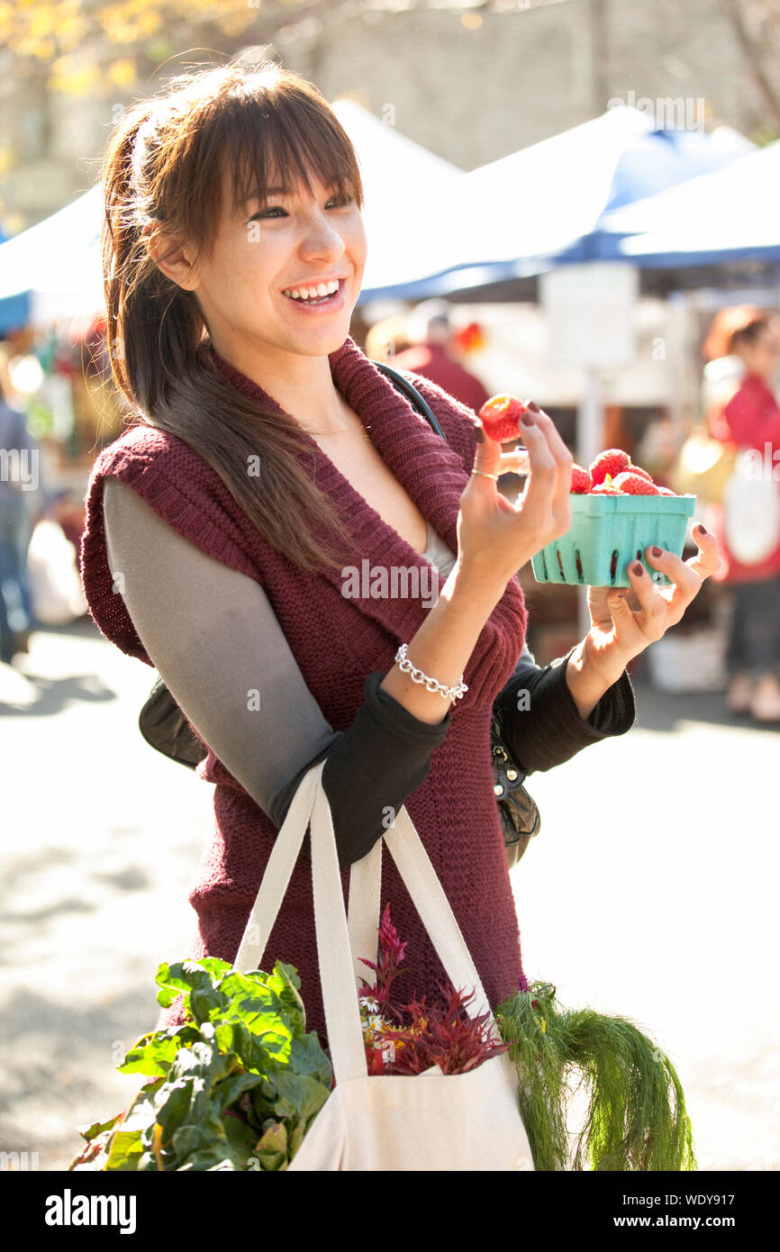 Beautiful, happy, young Hispanic woman eating fresh, organic strawberries fruit at farmers market. Healthy lifestyles. Stock Photo