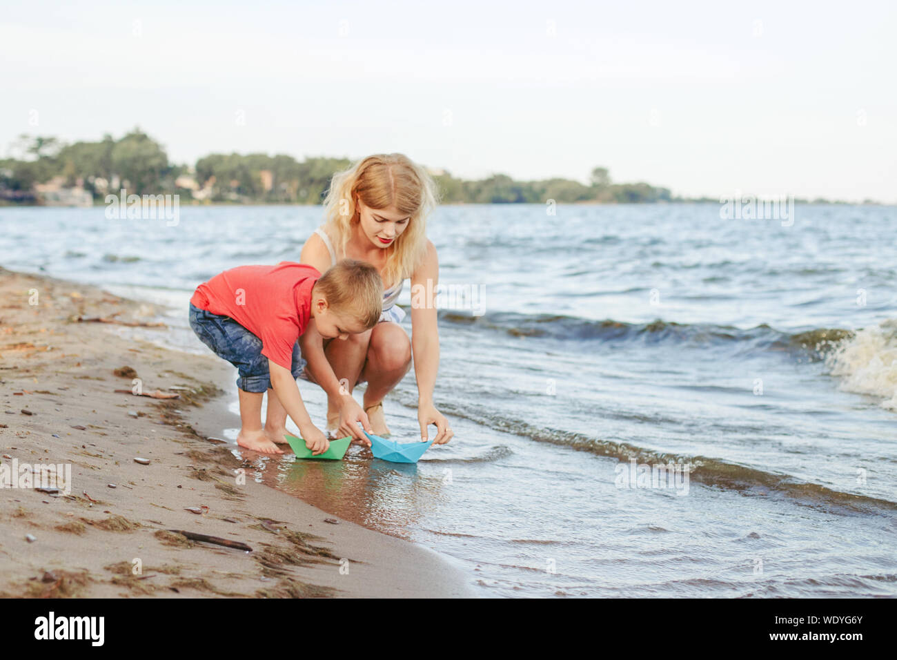 Caucasian mother with toddler son child putting paper boats in water together on lake shore. Kid playing on beach. Outdoor family summer activity. Hap Stock Photo