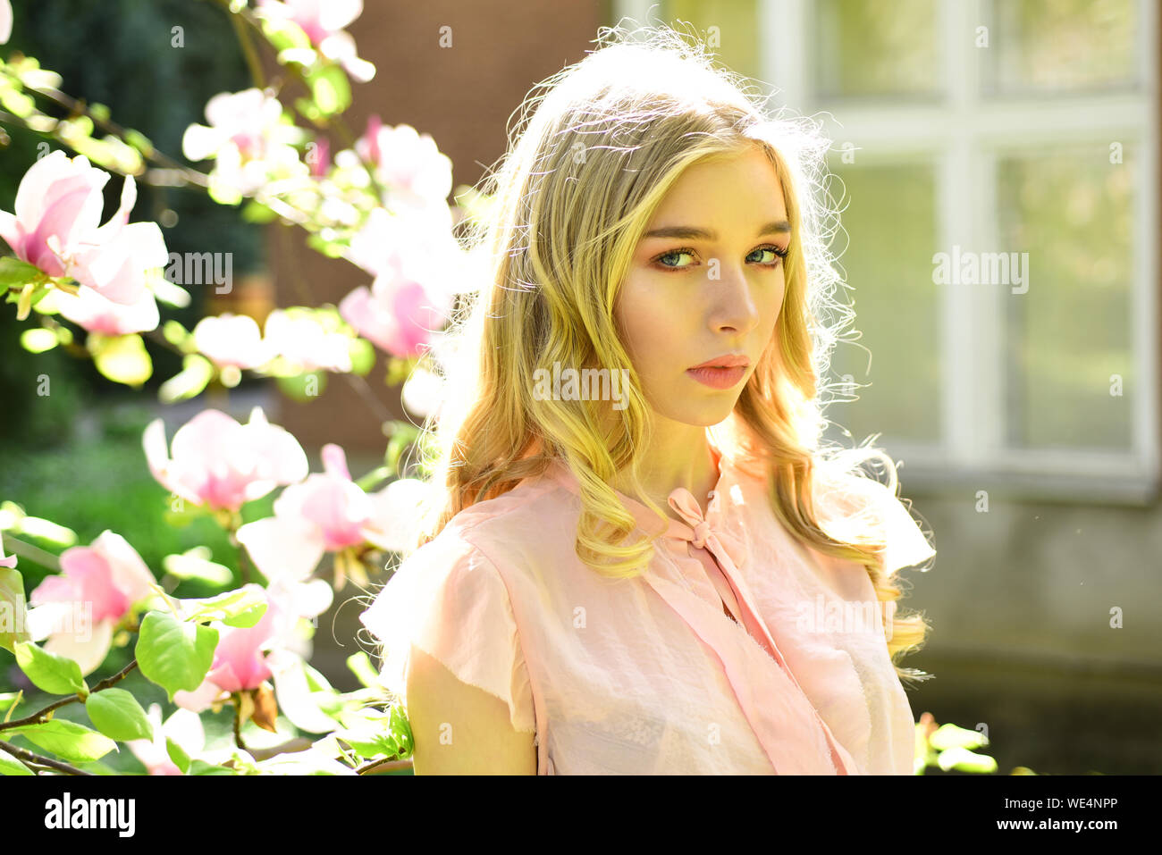 Attractive curly-haired woman walks in spring garden, enjoying the scent of flowering trees. Portrait of charming blond girl in front of blooming Stock Photo
