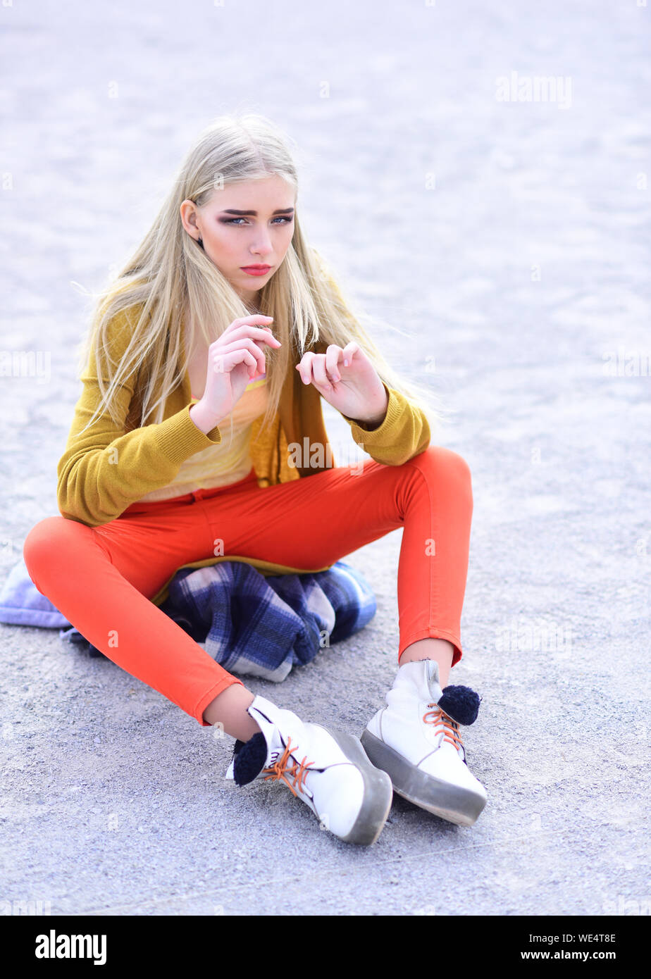 Blond model with thick eyebrows and blushed cheeks sitting on sandy road. Girl in bright outfit posing outdoors. Street fashion concept Stock Photo