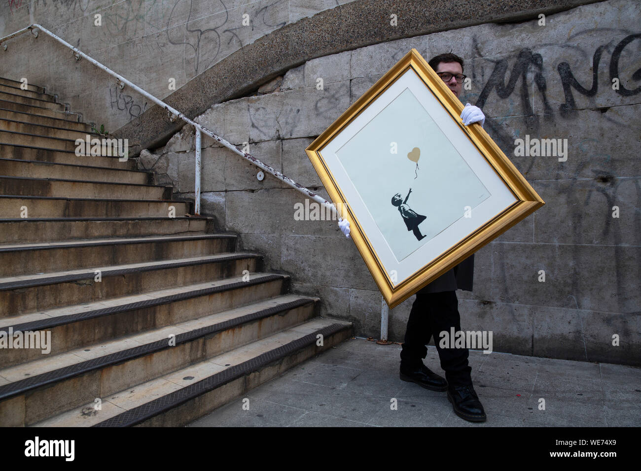 London, UK. 30th Aug, 2019. A Christie's employee poses with artworks 'Girl with balloon - Colour AP (Gold)' by anonymous street artist Banksy at the Southbank Centre in London Friday, August 30, 2019. The Southbank Centre was near one of the original locations the artwork appeared in London. The screen prints will be on sale during an online only auction titled 'Banksy : I can't believe you morons buy this sh*t' presented by Christie's auction house between 11-24 September 2019. Photograph Credit: Luke MacGregor/Alamy Live News Stock Photo