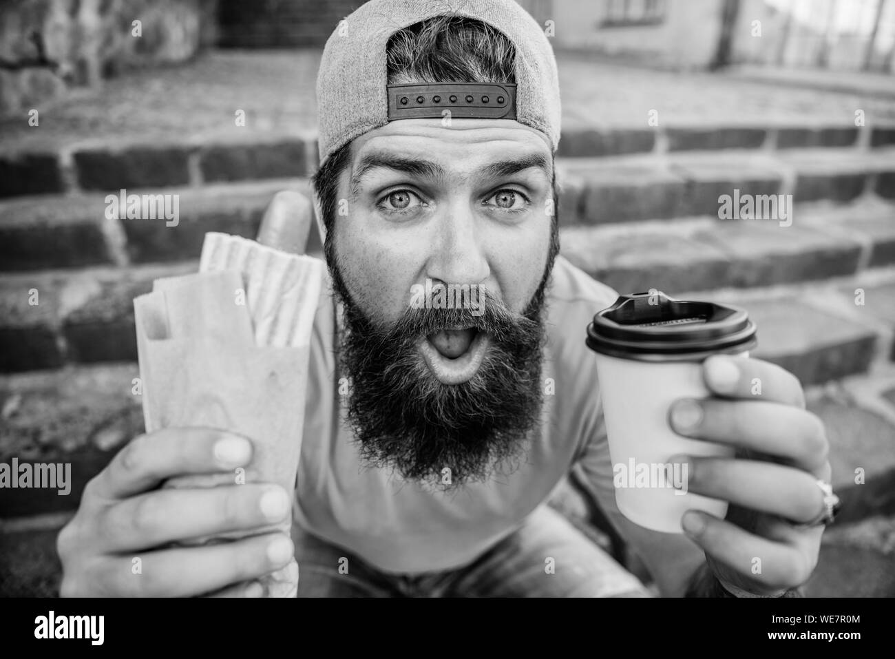 Urban lifestyle nutrition. Junk food. Carefree hipster eat junk food while sit stairs. Snack for good mood. Guy eating hot dog. Street food concept. Man bearded eat tasty sausage and drink paper cup. Stock Photo