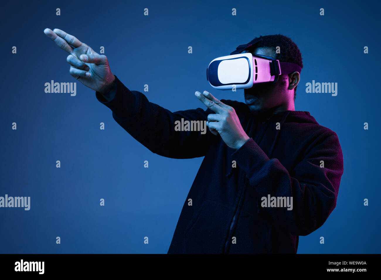 Young african-american man in VR-glasses in neon on blue background. Male portrait. Concept of human emotions, facial expression, modern gadgets and technologies. Pointing, shooting while gameplay. Stock Photo