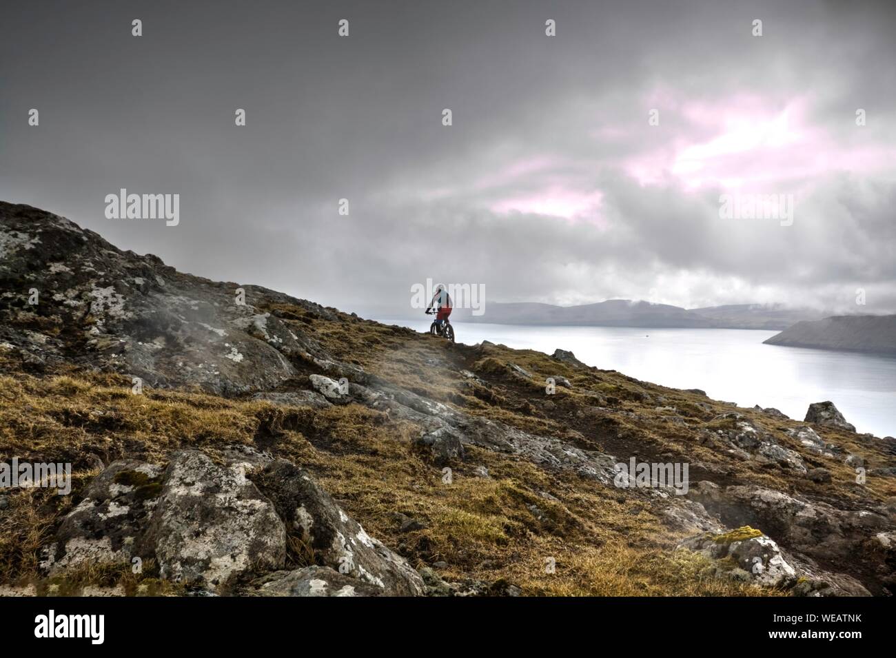 Person Mountain Biking On Rocky Cliff By Lake Against Cloudy Sky Stock Photo