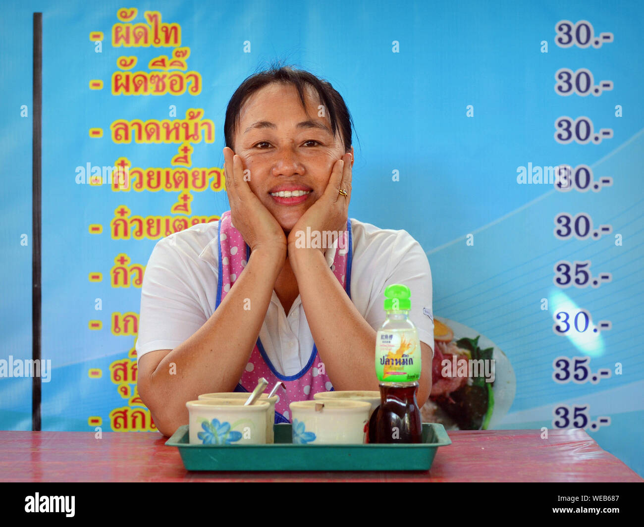 Thai female cook and co-owner of a local restaurant poses in front of the menu/price list and smiles for the camera. Stock Photo