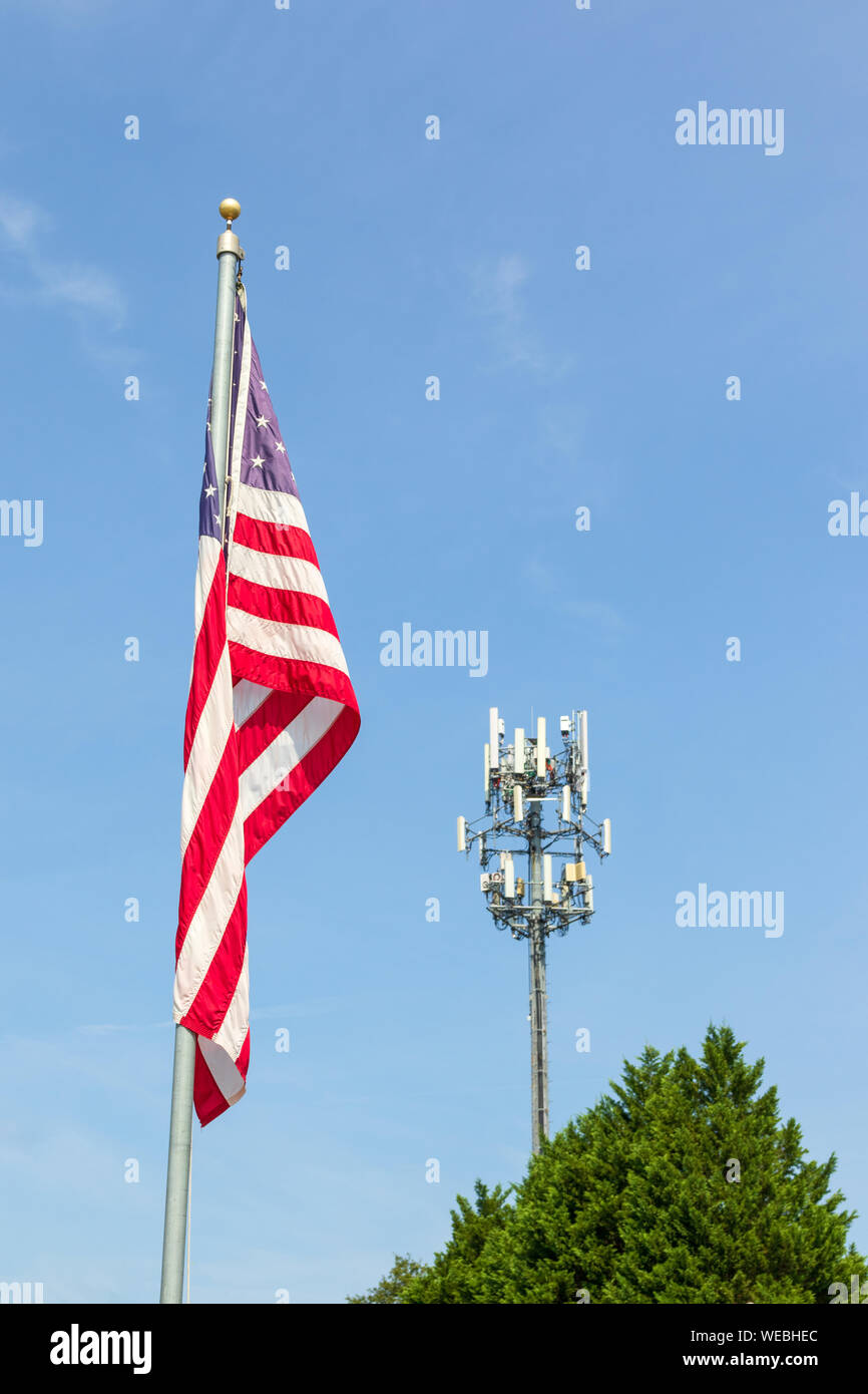 An American flag on flag pole juxtaposed to a cell phone tower and the peak of an evergreen tree.  Blue sky background.  Copy space. Stock Photo