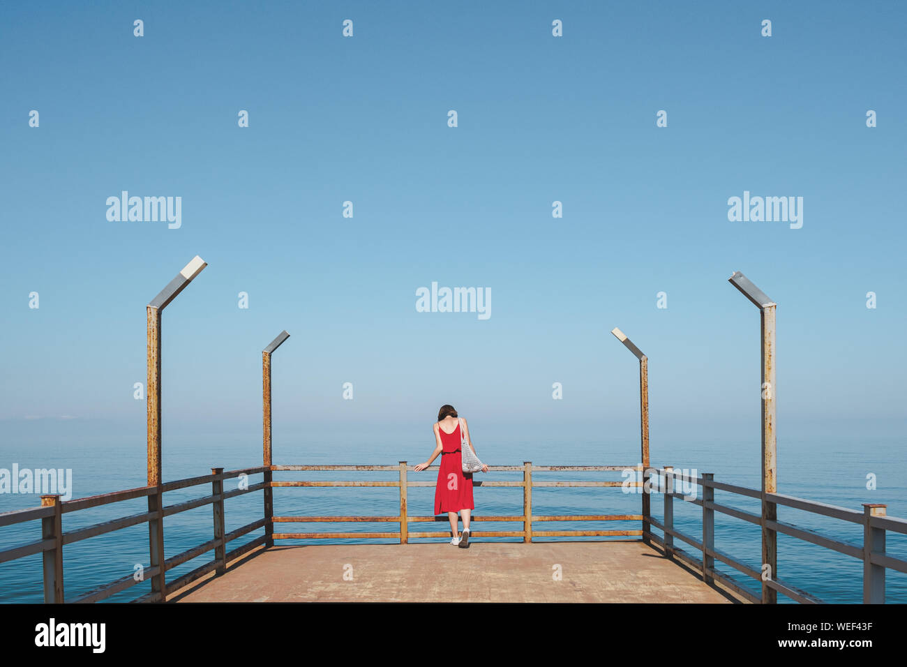 Standing on the pier by the beautiful blue sea. Woman in red dress ...