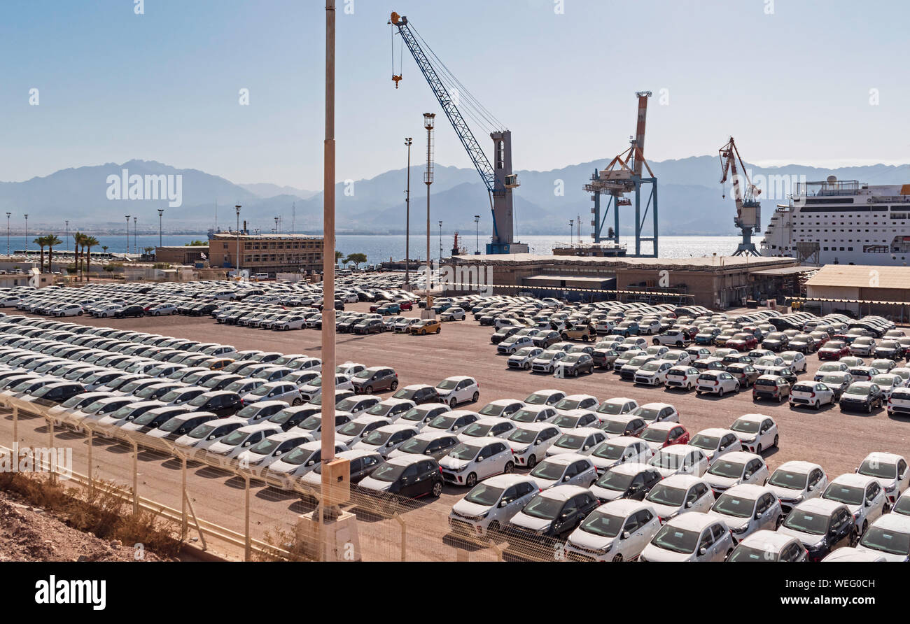 a sea of new cars in front of the docks of the port of eilat in israel with a cruise ship, cranes and the mountains of Jordan in the background Stock Photo