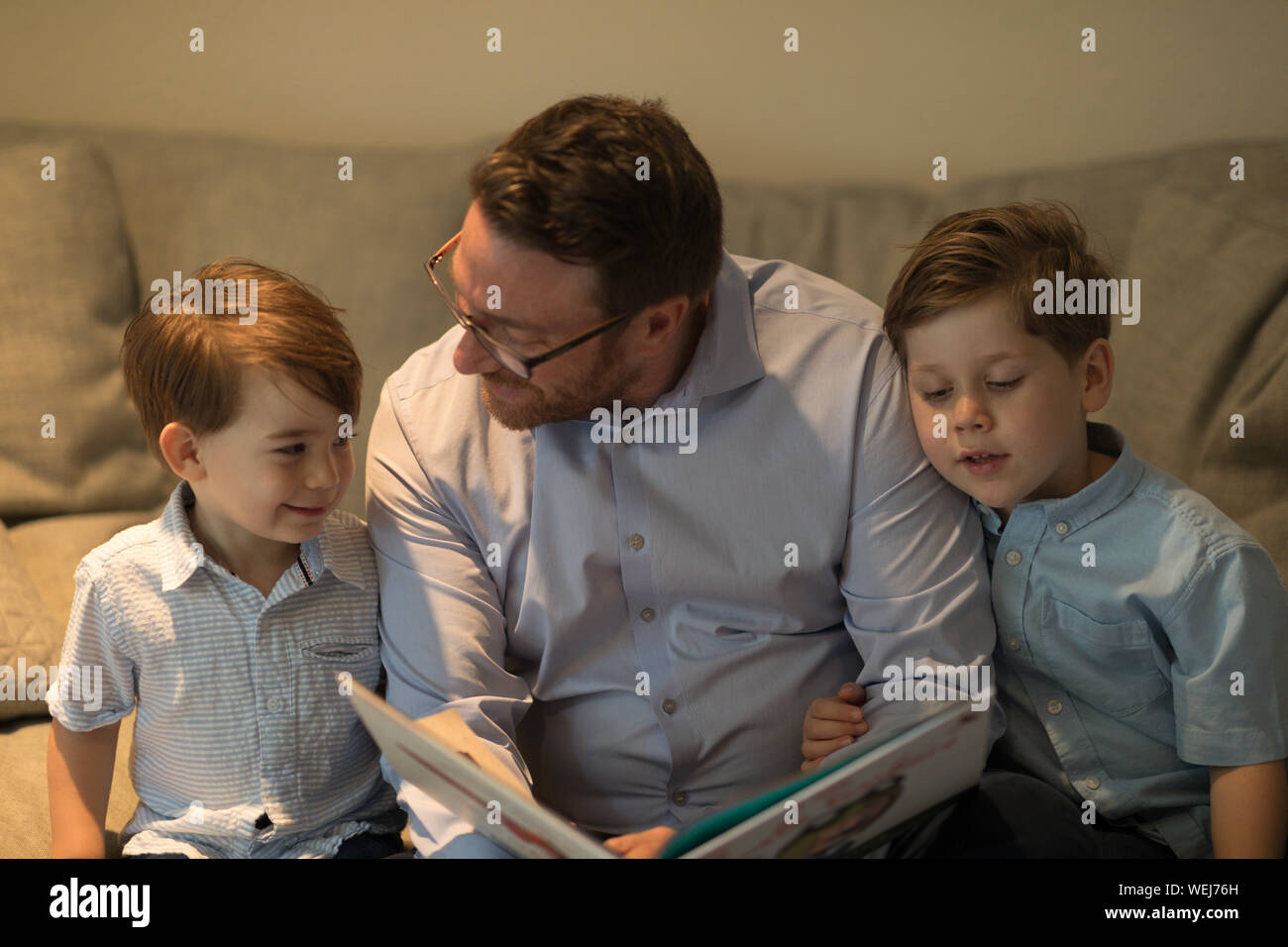 Father with two sons, 2 and 4, looking at book on sofa Stock Photo