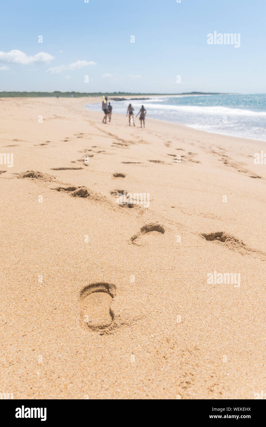 Footprints along the sand coast, a group of four people are hanging out in the beach, the ocean has just a single wave, in the horizon there are some Stock Photo