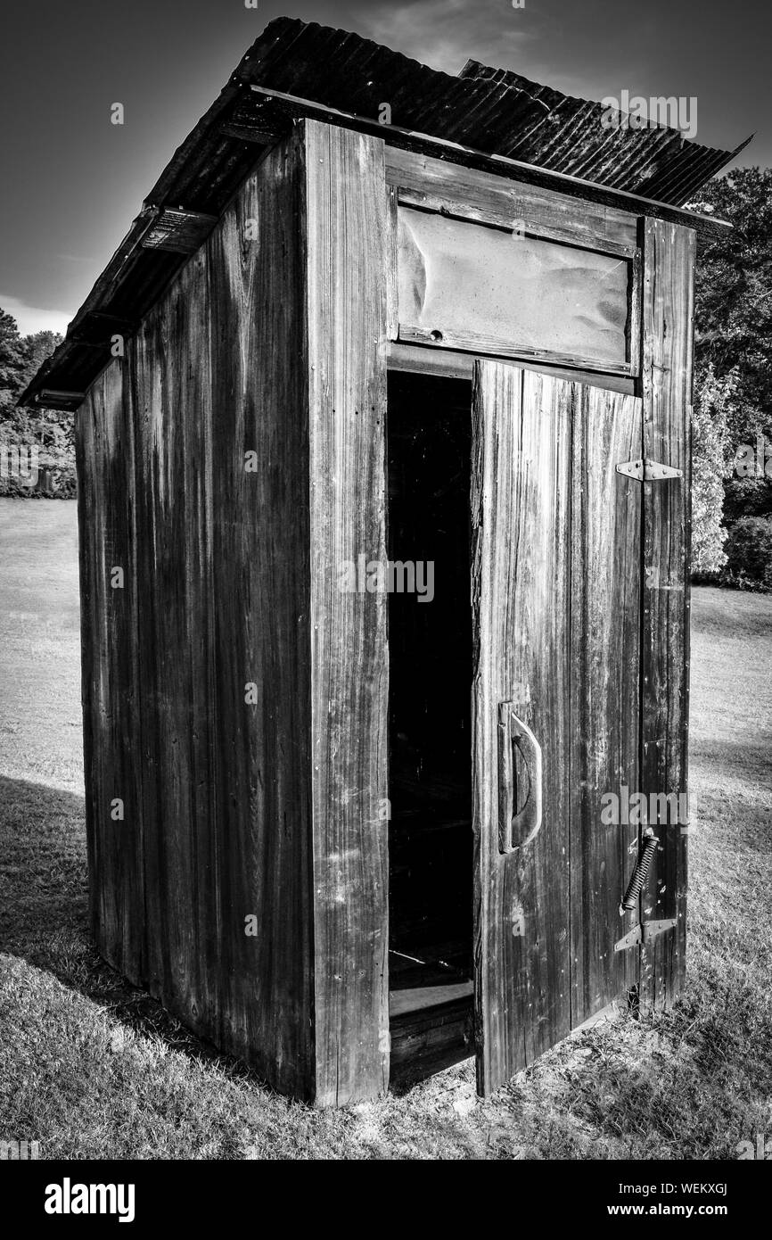 Old,  weathered out-house, or toliet, with door ajar with rusty roof on a hillside field with trees in the Southeastern USA , in black and white Stock Photo
