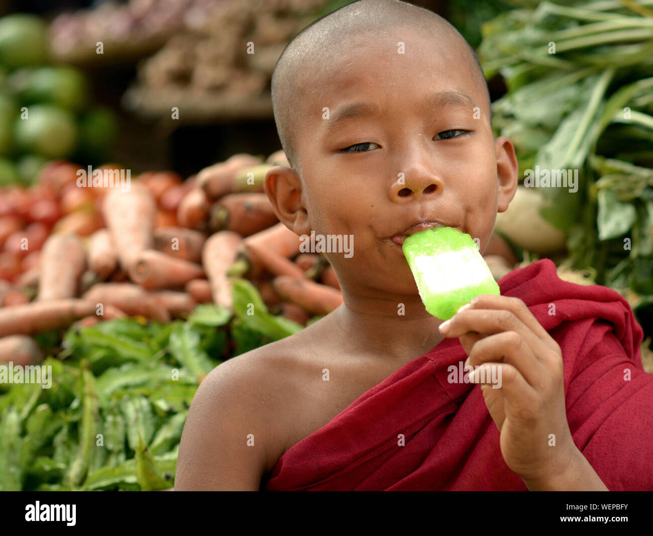 Burmese Buddhist boy monk eats a cheap ice-cream bar at the local produce market. Stock Photo