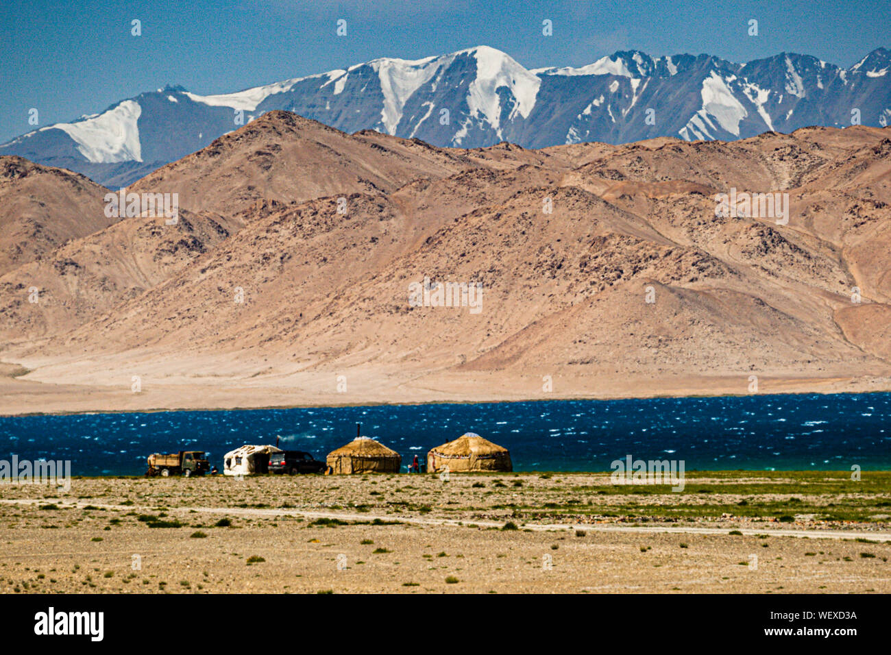 The yurts of Kyrgyz nomads sit beside the parked vehicles on the shore of Lake Karakul in Tajikistan, the snow-capped peaks of the Pamir mirrored in the deep blue-turquoise water. Stock Photo