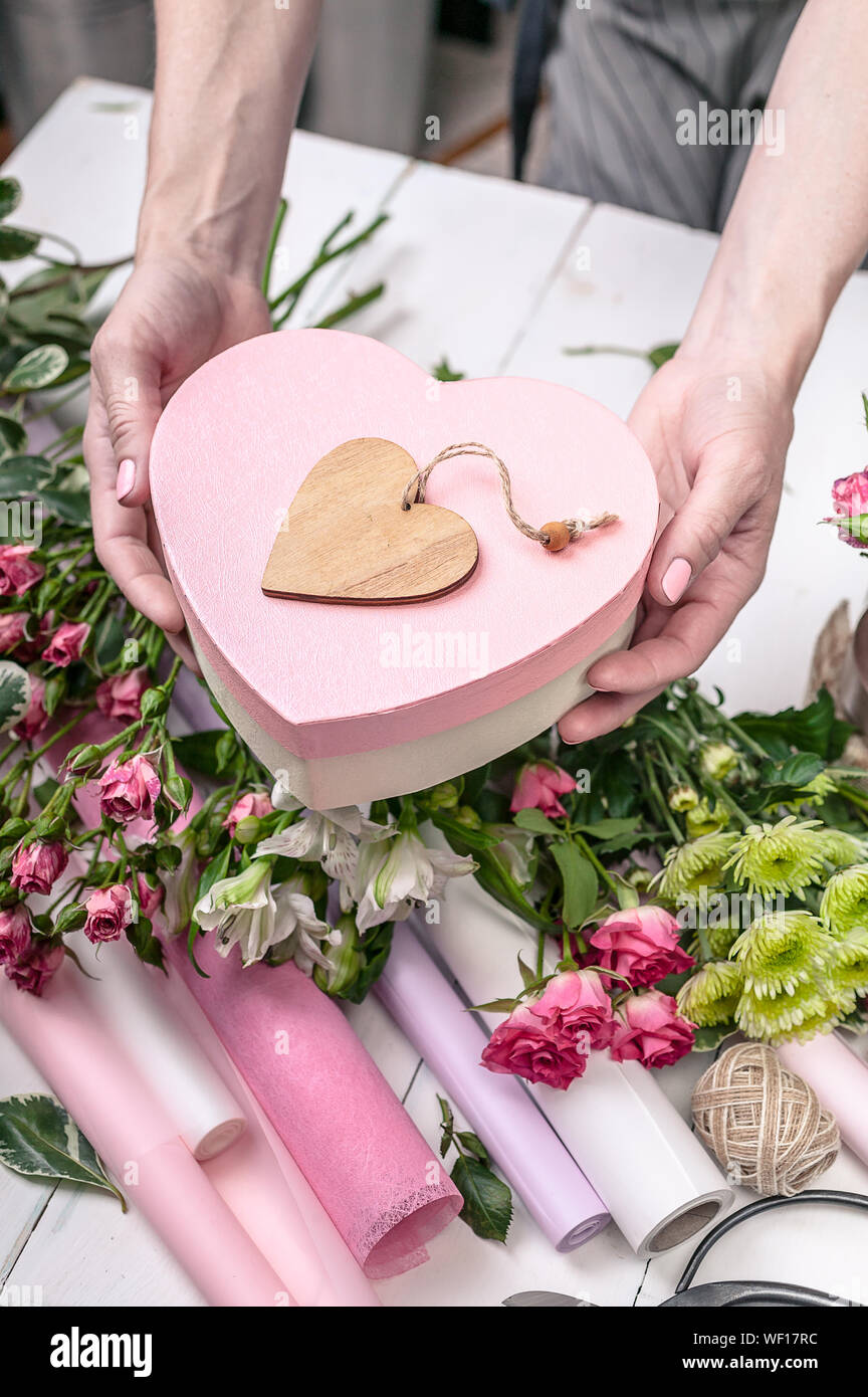 Female hands holding a gift box in the shape of a heart. Florist's workplace: flowers, accessories, tools. Stock Photo