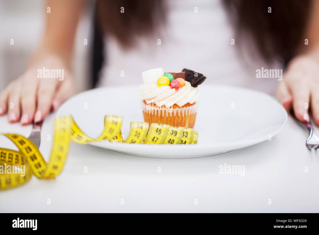 Young dieting woman sitting in front of plate with delicious cream tart cakes, looking at forbidden food with unhappy and hungry expression, studio Stock Photo