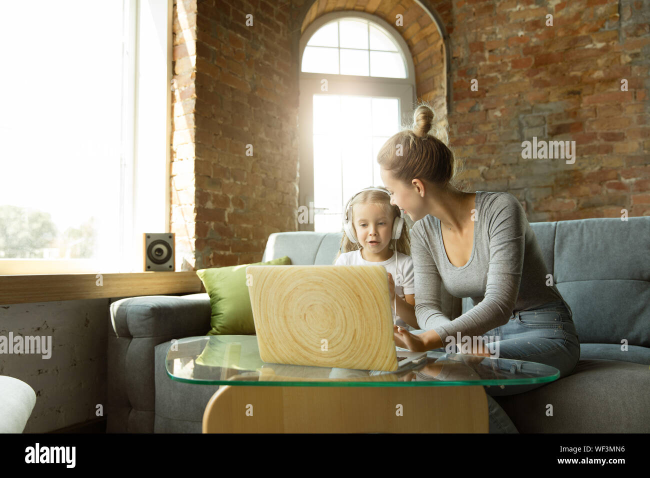 Female caucasian teacher and little girl, or mom and daughter. Homeschooling. Sitting on the sofa and using laptop for knowledges getting while lesson is. Education, school, studying concept. Stock Photo