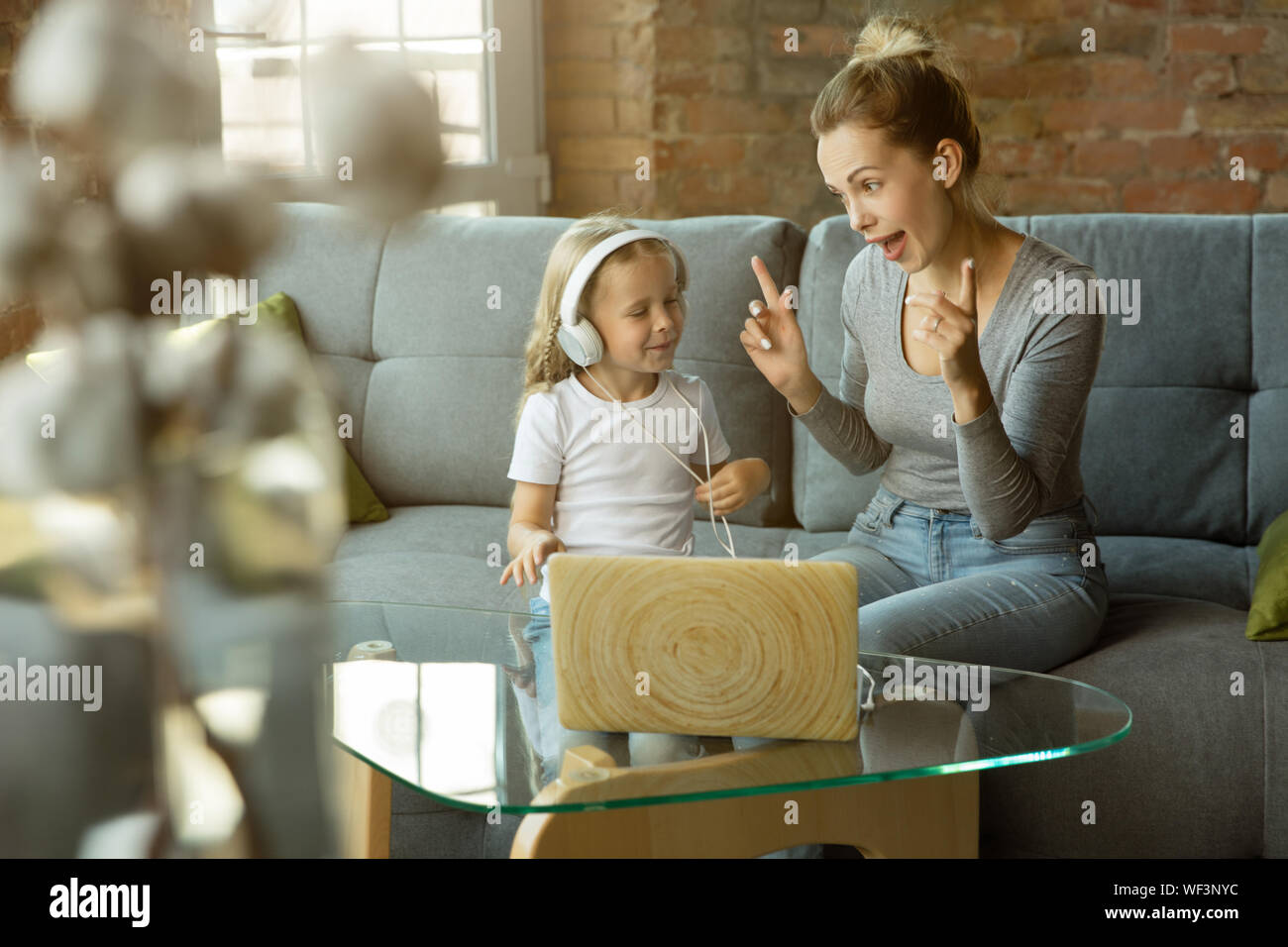 Female caucasian teacher and little girl, or mom and daughter. Homeschooling. Sitting on the sofa and using laptop for knowledges getting while lesson is. Education, school, studying concept. Stock Photo