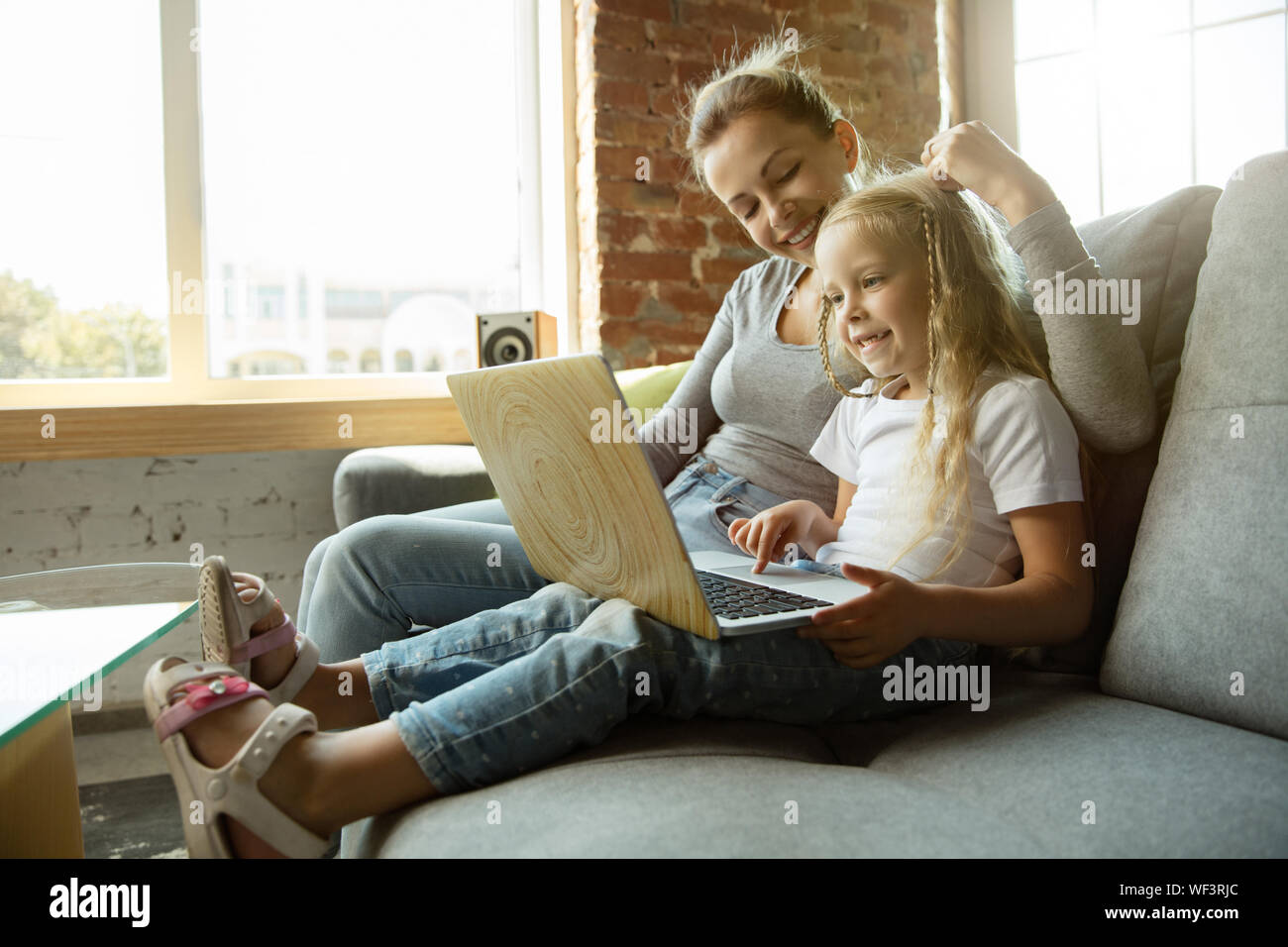 Female caucasian teacher and little girl, or mom and daughter. Homeschooling. Sitting on the sofa and using laptop for knowledges getting while lesson is. Education, school, studying concept. Stock Photo