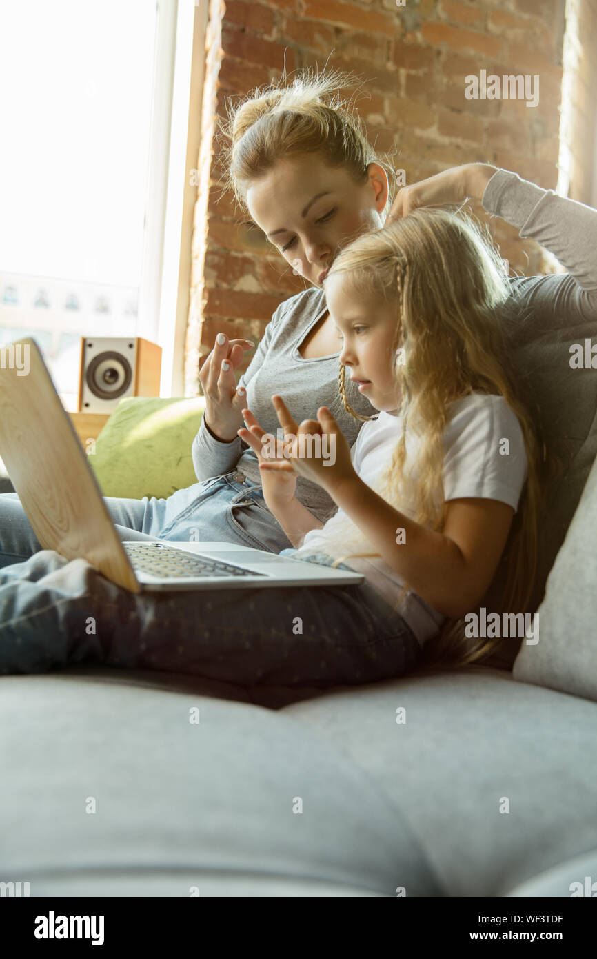 Female caucasian teacher and little girl, or mom and daughter. Homeschooling. Sitting on the sofa and using laptop for knowledges getting while lesson is. Education, school, studying concept. Stock Photo