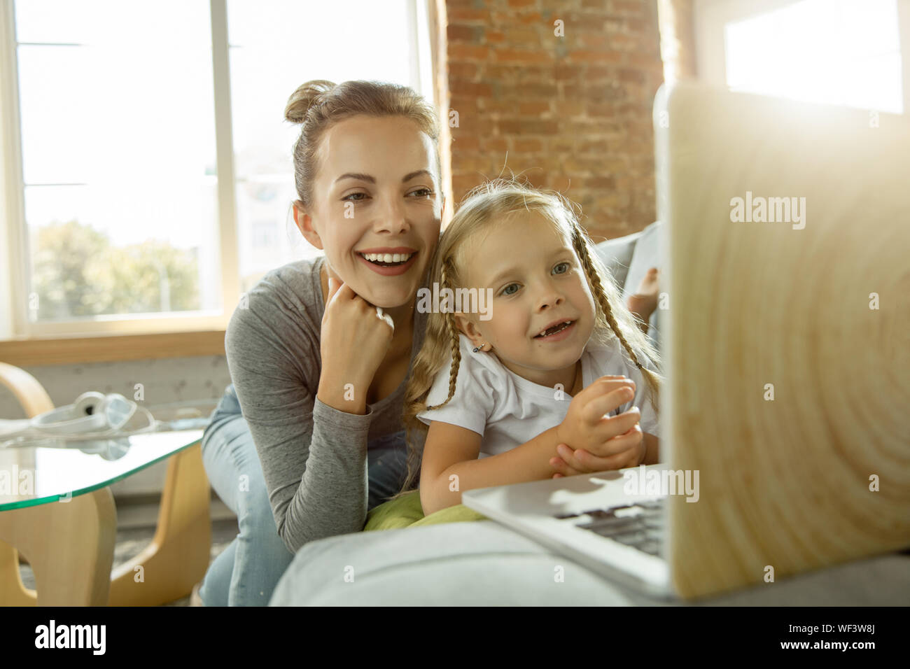 Female caucasian teacher and little girl, or mom and daughter. Homeschooling. Sitting on the sofa and using laptop for knowledges getting while lesson is. Education, school, studying concept. Stock Photo