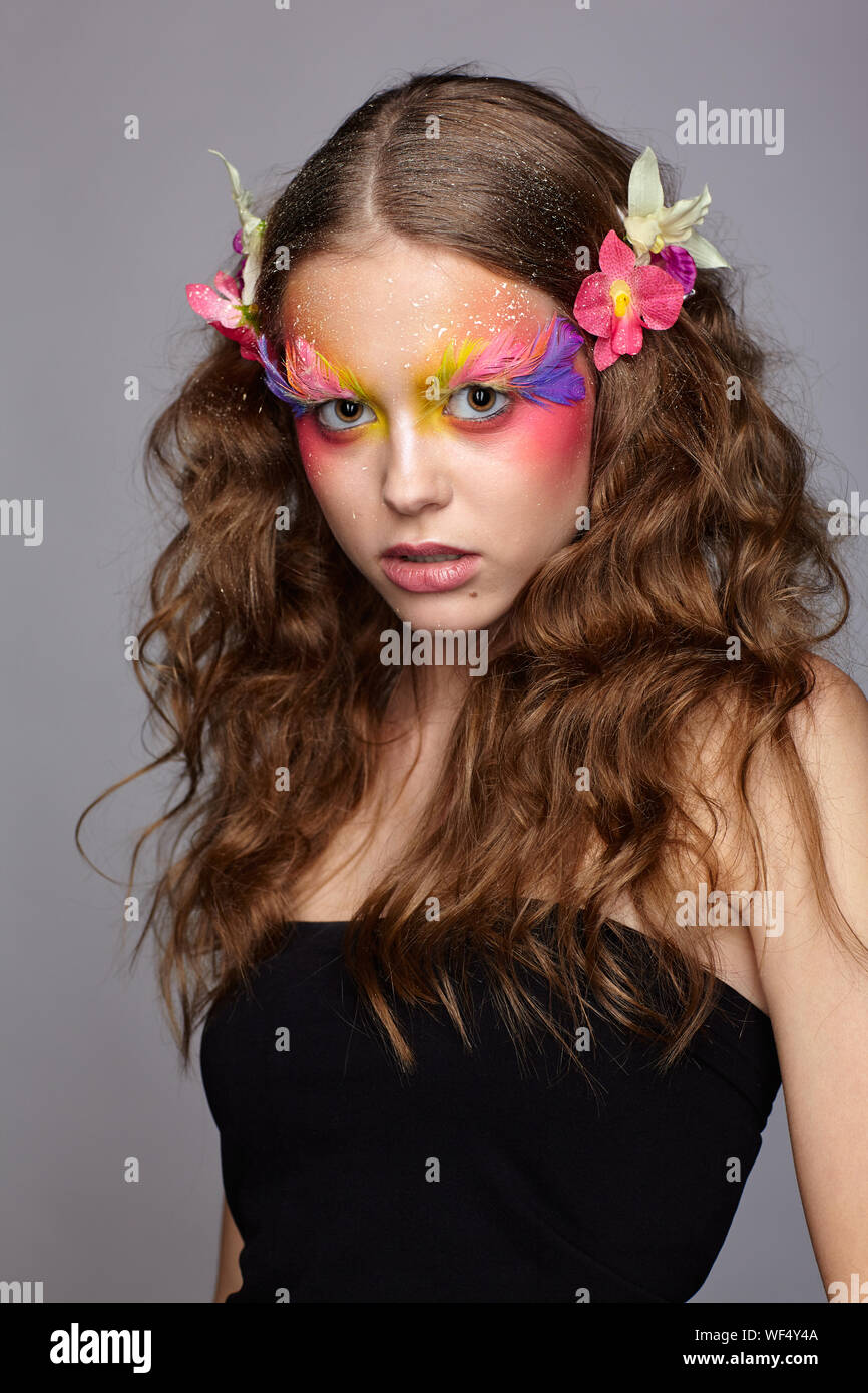 Portrait of teen girl with orchid flower in wavy hair. Young female with unusual stylish make-up and false fashion feather eyelashes. Stock Photo