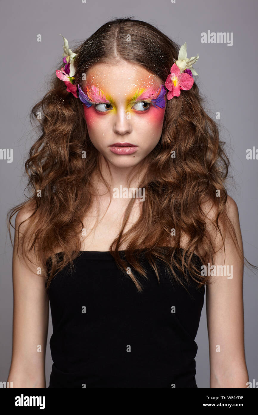 Portrait of teen girl with orchid flower in wavy hair. Young female with unusual stylish make-up and false fashion feather eyelashes. Stock Photo