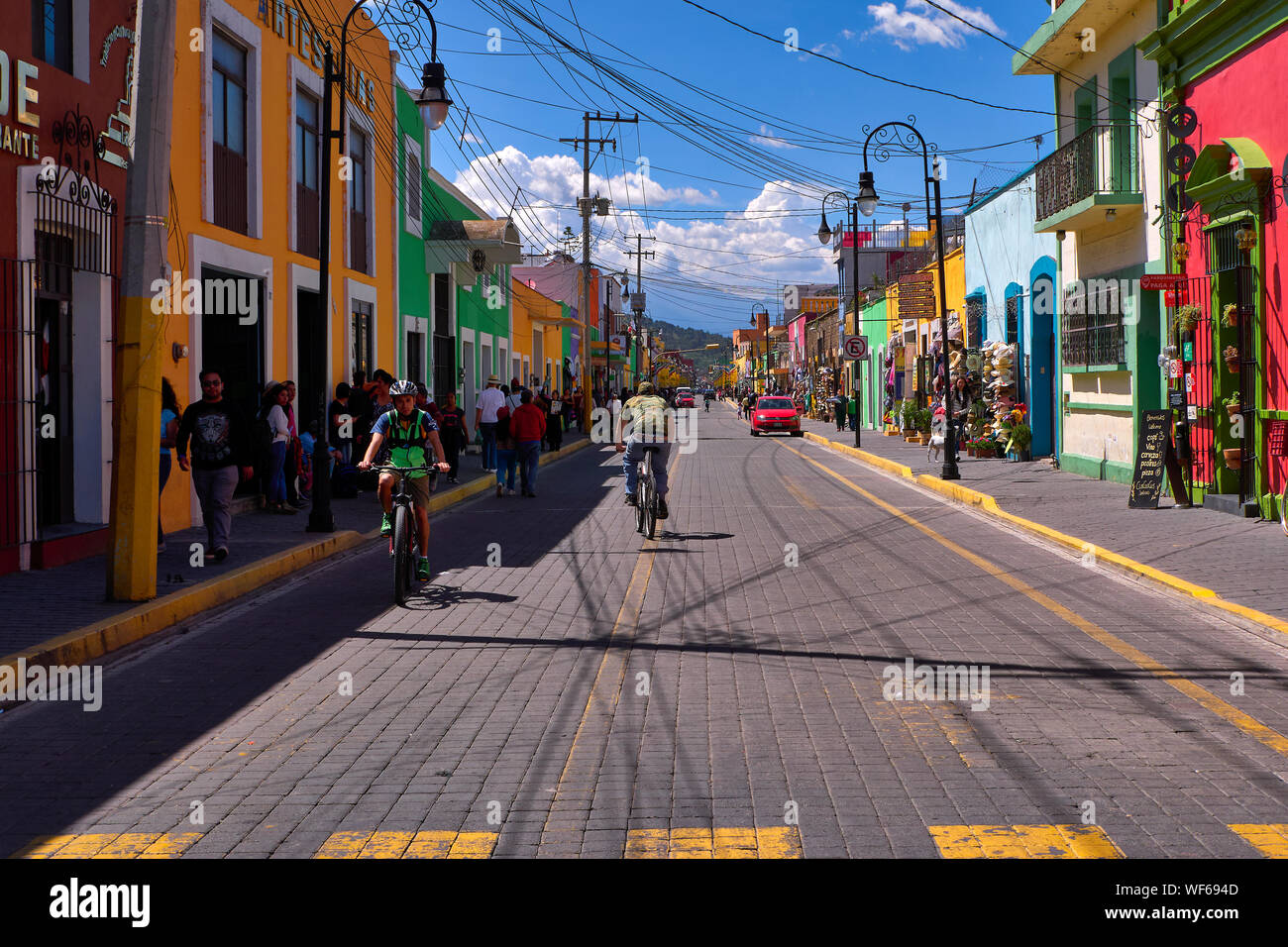San Pedro Cholula, Mexico, September 30, 2019 - Beautiful Avenida Morelos street with 2 cyclists and tourists at sunny day, traditional clothing and souvenir shops with vibrant colored facades. Stock Photo
