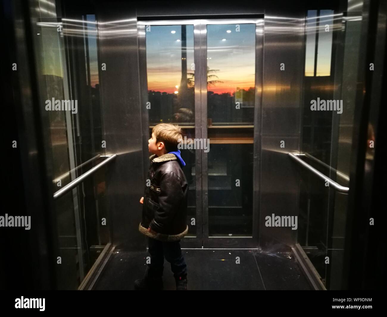 Side View Of Boy Standing In Glass Elevator During Sunset Stock Photo