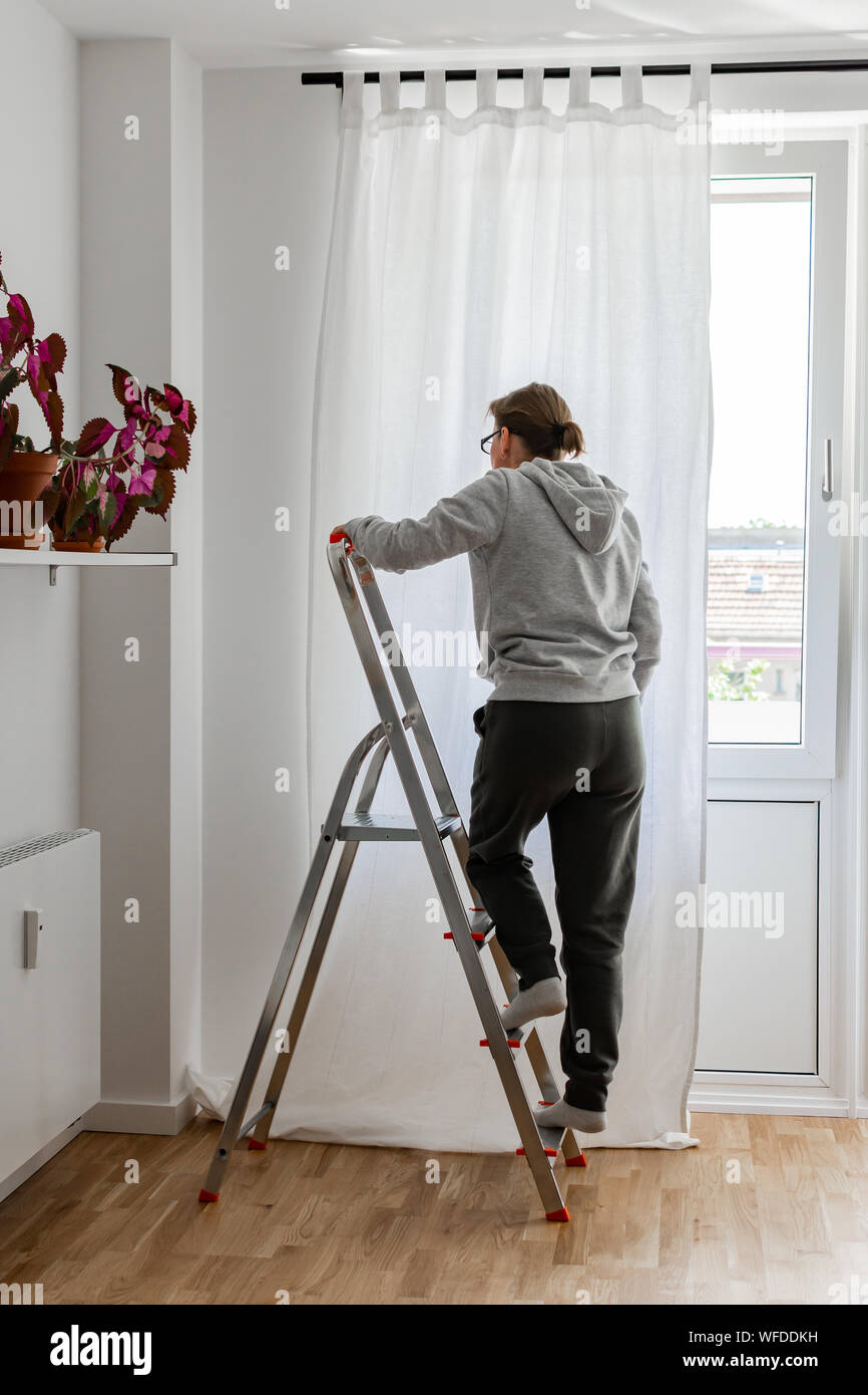 Woman Stands On A Stepladder Near The Window And Hangs White