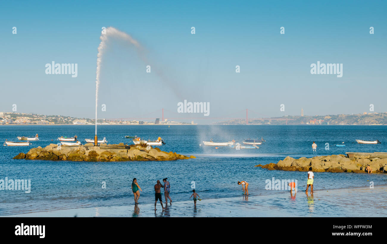 Lisbon, Portugal - August 30, 2019: People on a beach near Lisbon with a geyser overlooking the iconic 25 April Bridge and Rei Cristo statue Stock Photo