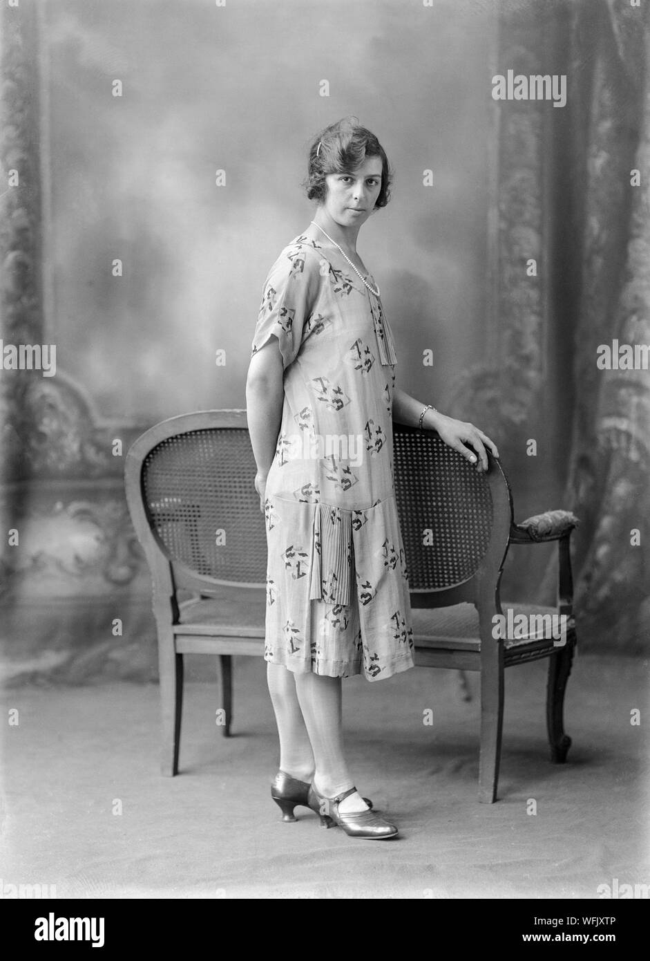 Early twentieth century black and white vintage photograph, taken in a photographic studio, showing a young woman, in a dress typical of the fashion of the period, posing for the camera whilst standing. Stock Photo