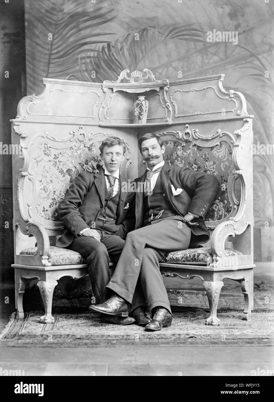A vintage late Victorian or early Edwardian black and white photograph showing two men, seated, posing in a photographic studio. One man has a very large moustache. The pose is very formal, but also quite relaxed. Stock Photo