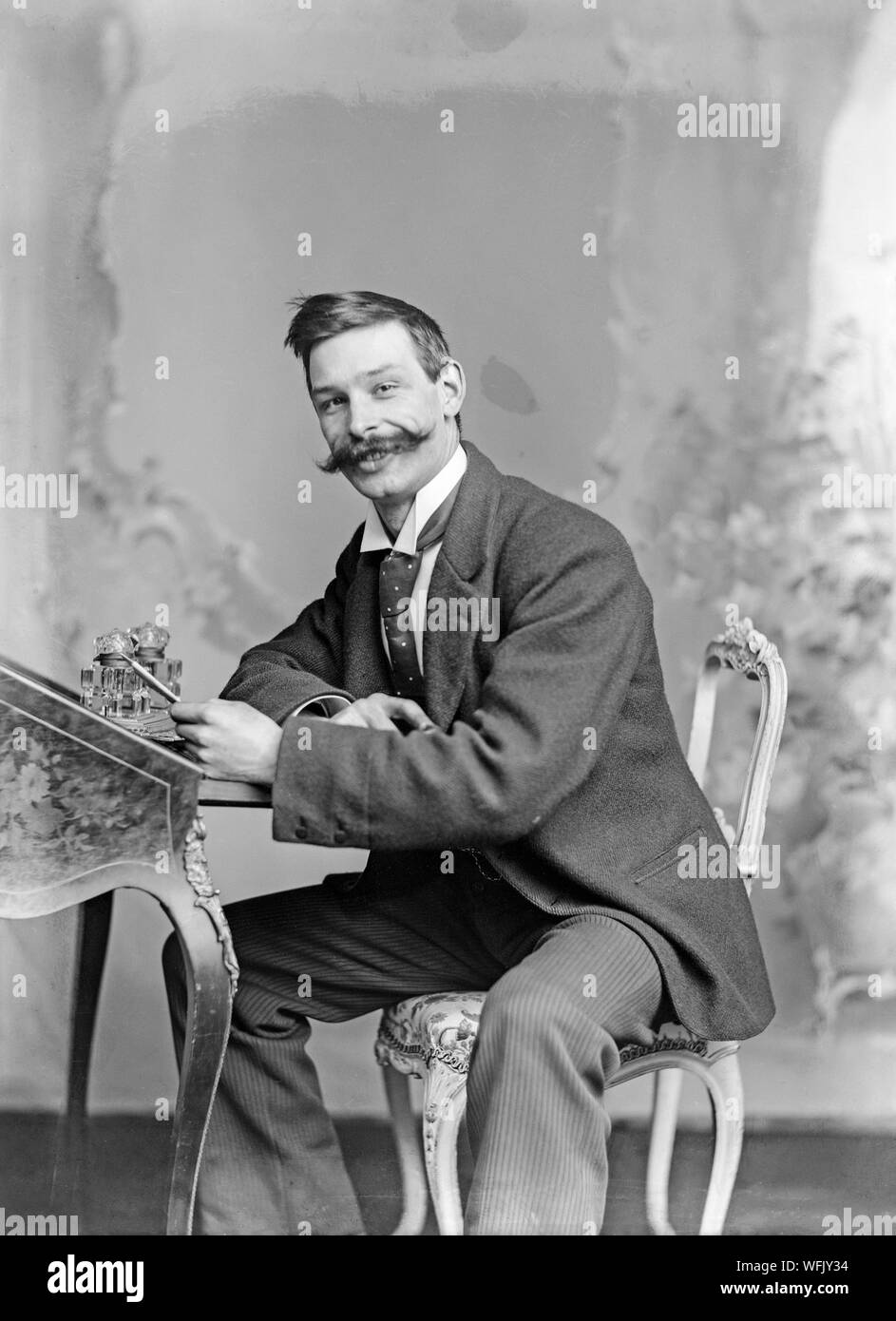 A vintage late Victorian or early Edwardian black and white photograph showing a young man with a very big moustache sitting at a writing desk with a pen in his hand, whilst he looks towards the camera smiling. Stock Photo