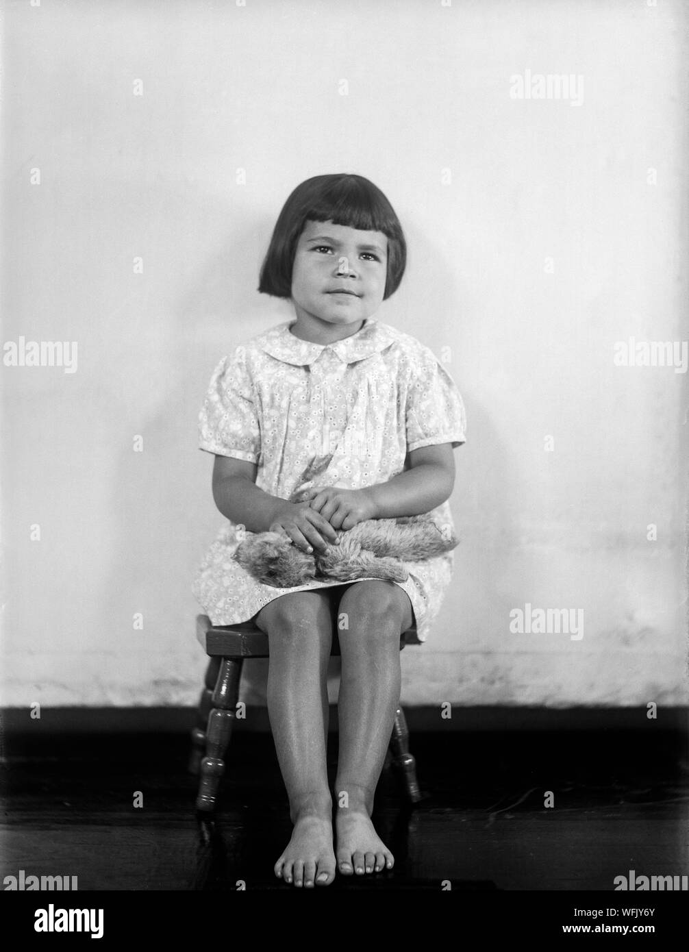 A vintage late Victorian or early Edwardian black and white photograph showing a young girl with a short bobbed haircut, sitting in a chair facing the camera. She is barefoot, and holding a toy teddy bear. Stock Photo