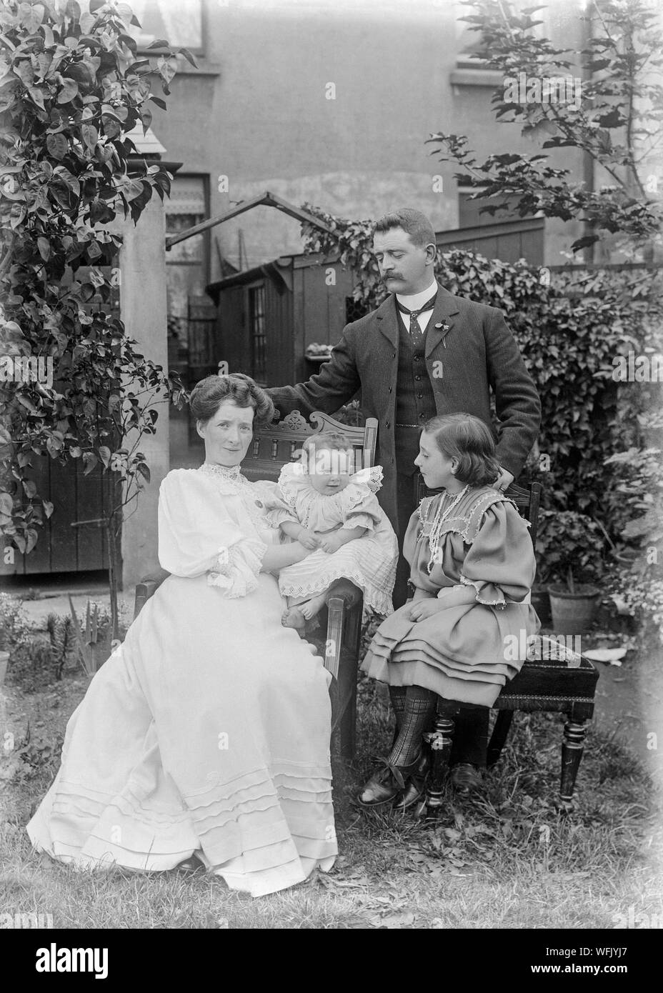 A vintage late Victorian or early Edwardian black and white photograph showing a family group of a father, mother, and two daughters, posing for the camera outdoors, in a suburban garden in England. They are wearing typical fashion of the era. Stock Photo