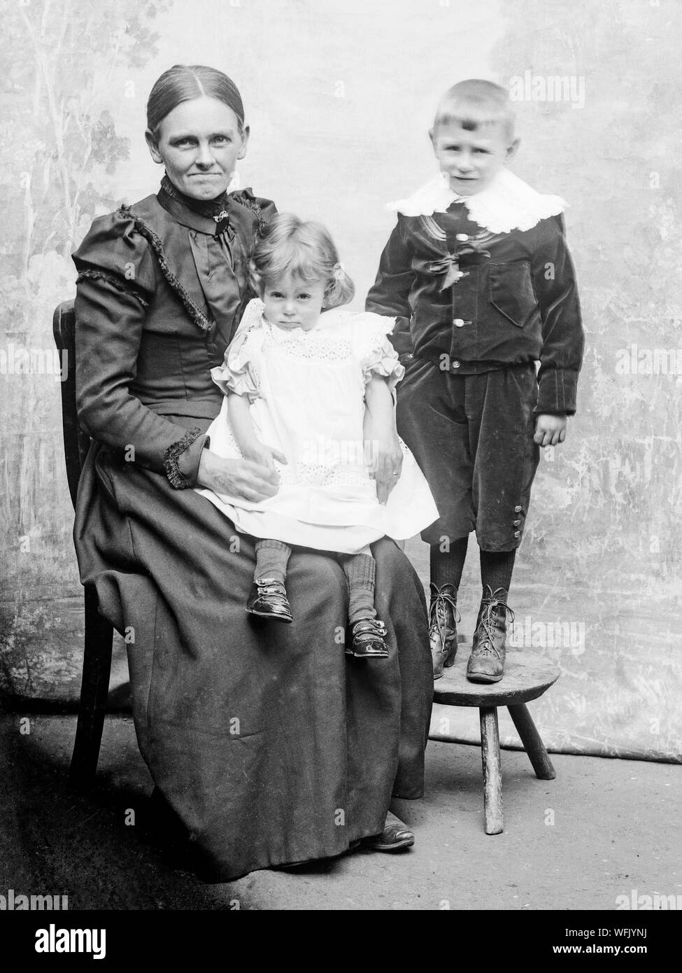 A vintage late Victorian or early Edwardian black and white photograph showing  a mother posing, sitting down, in a studio, with her bay daughter and young son. They show typical fashion of the period. Stock Photo
