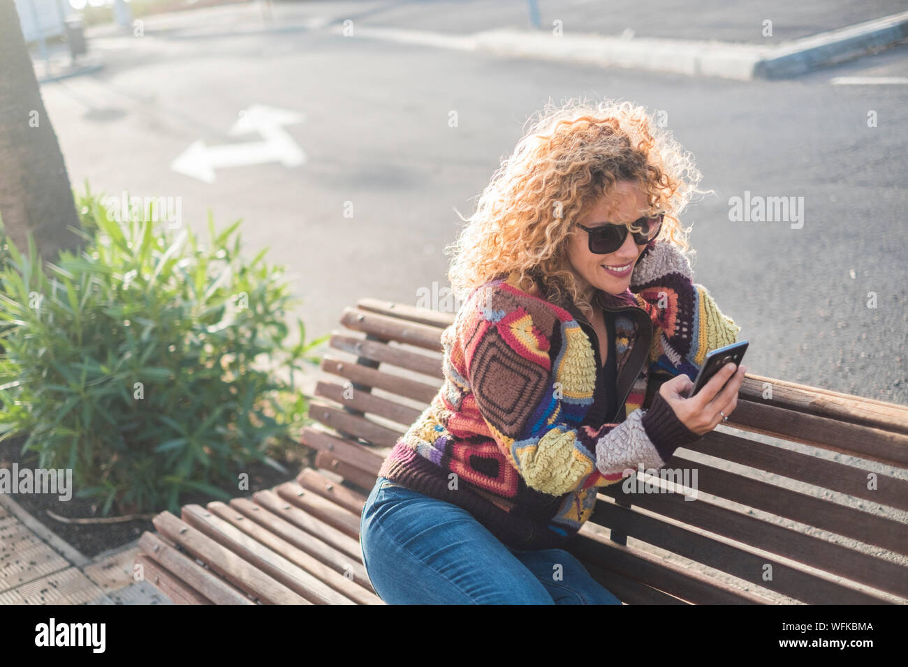 Woman Alone In The Street Sitting On A Bench Of The Park Using Her
