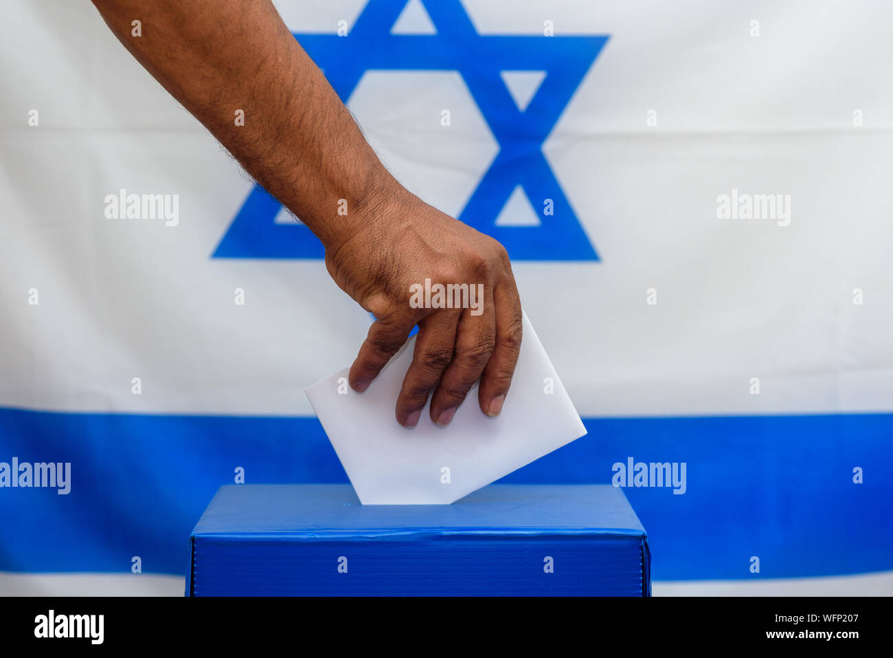 Israeli man putting a ballot in a ballot box on election day. Close up of hand with white votes paper on Israel flag background. Mockup, space for text. Stock Photo