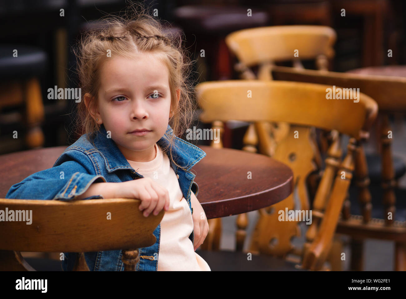 Fashionable little schoolgirl sitting at table of summer terrace of restaurant. She's melancholing. Text space Stock Photo