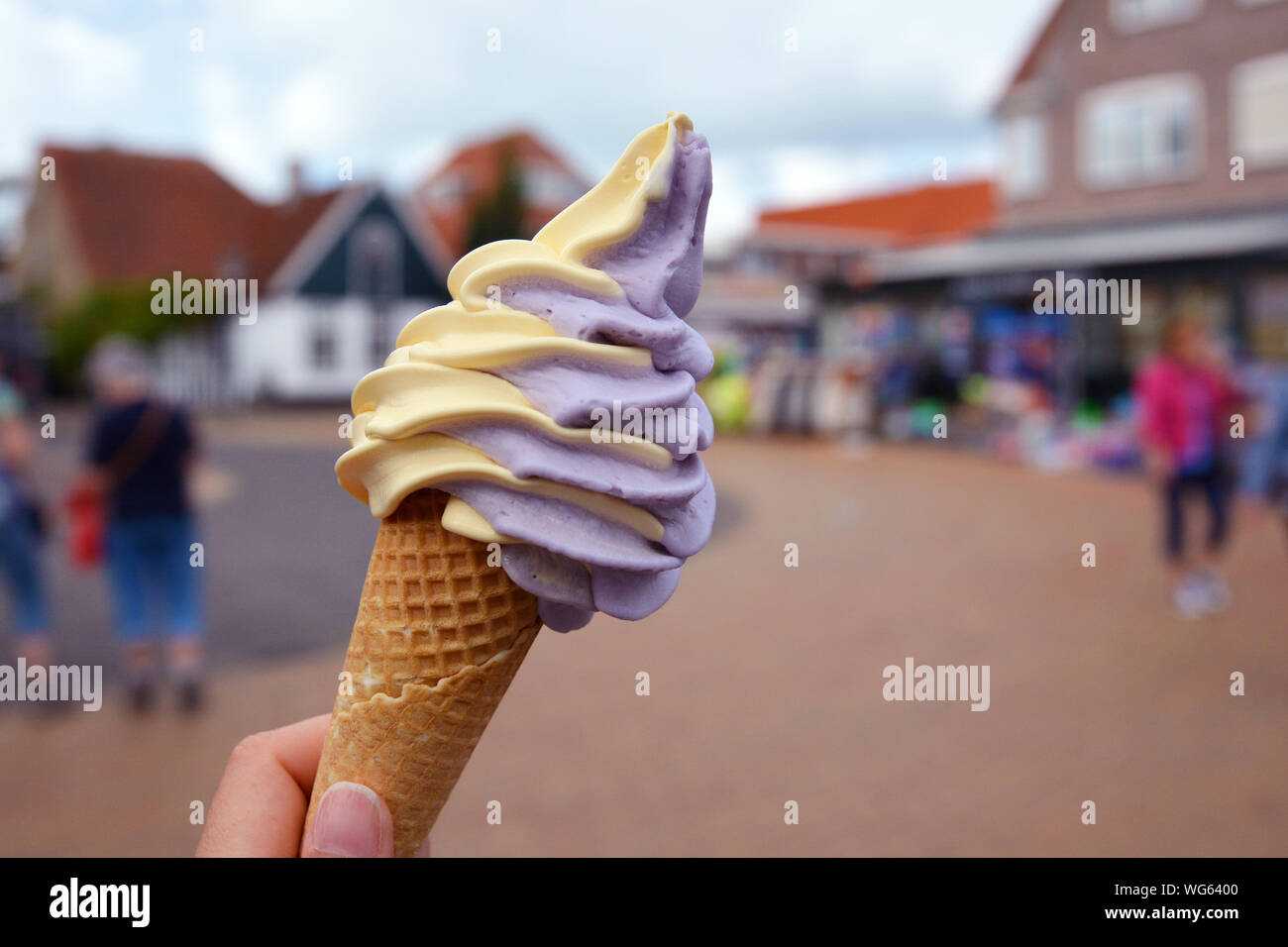 Close up of hand holding cone with mixed yellow and purple mango and berry soft serve ice cream with blurry city scene in background Stock Photo