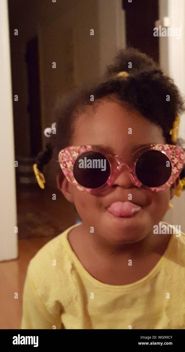 Close-up Of Little Girl Wearing Sunglasses While Sticking Out Tongue At Home Stock Photo
